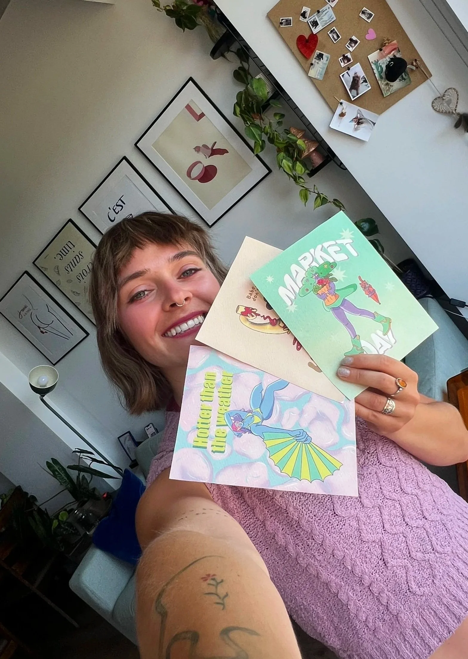 A young woman with short light brown hair and tattoos on her arm is smiling and holding four colorful greeting cards, with a background of framed artwork, a bulletin board with photos and decorations, plants, and a lamp.