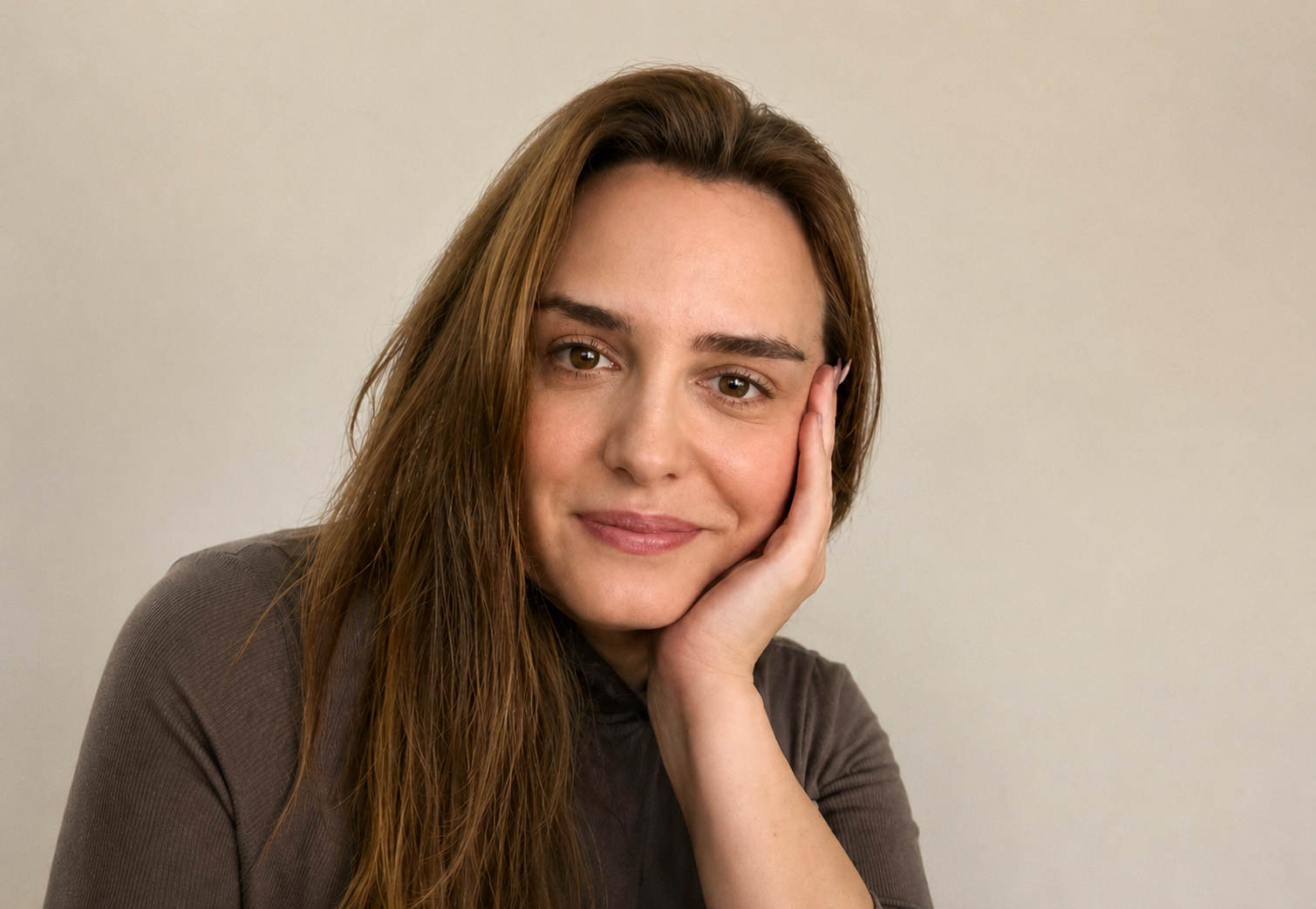 Junge Frau mit langen braunen Haaren, lächelnd, die ihr Gesicht mit Hand stützt, vor hellem Hintergrund.