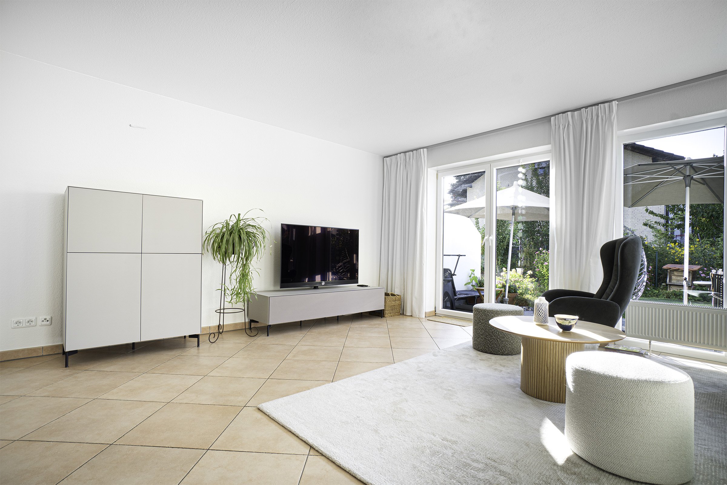 Ein helles, modernes Wohnzimmer mit großen Fenstern, weißen Vorhängen, einem Fernseher, einer schwarzen Sessel, einem weißen Teppich und Blick auf einen Garten mit Sonnenschirm und Gartenmöbeln.