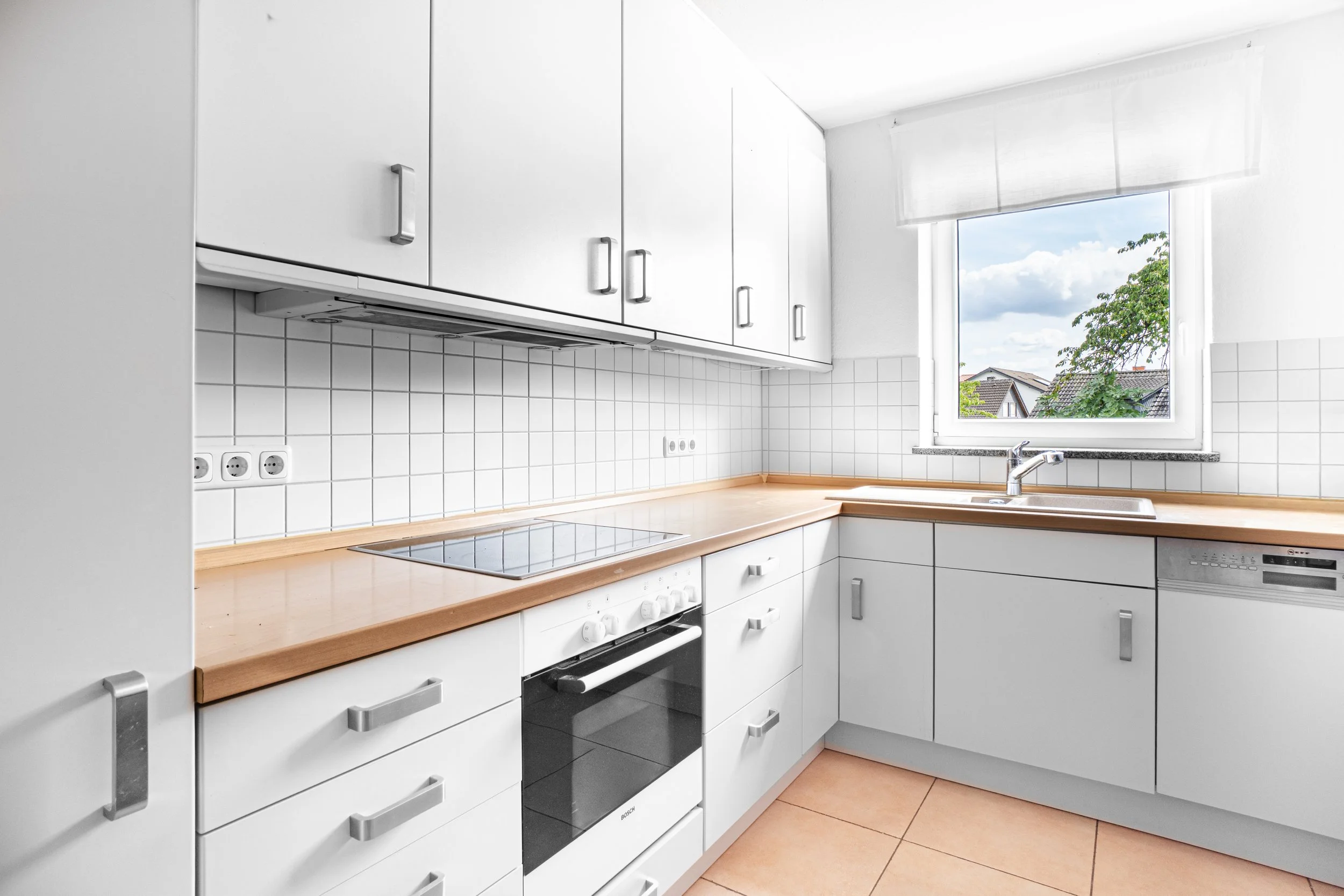 Küche mit weißen Schränken, Herd, Backofen, Spüle vor einem Fenster mit Blick auf Wohnhäuser und Bäume, helle Wandfliesen, Holztisch.