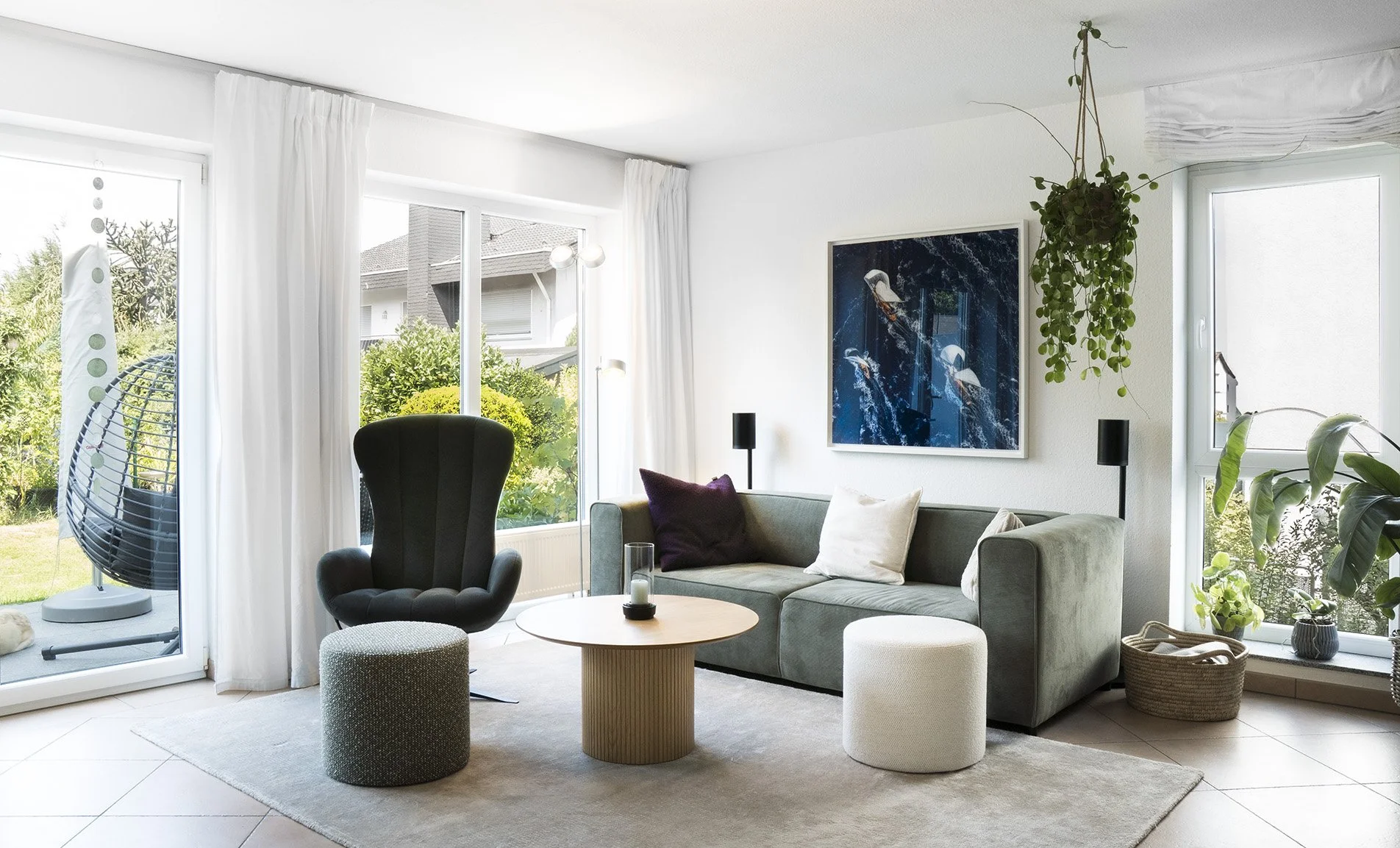 Helles Wohnzimmer mit großen Fenstern, grüner Couch, schwarzen Armstuhl, Trockenpflanze und Wandbild mit Wasser-Motiv.