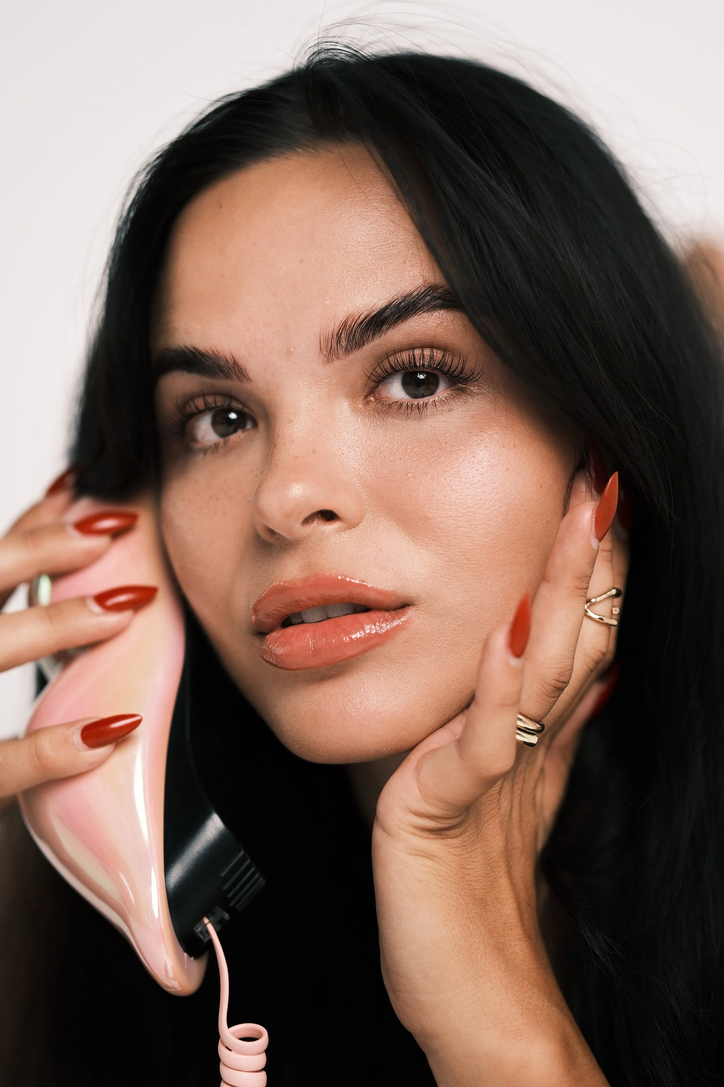 A woman with long black hair holding a vintage pink rotary telephone to her ear, touching her face with her free hand, and gazing into the distance.