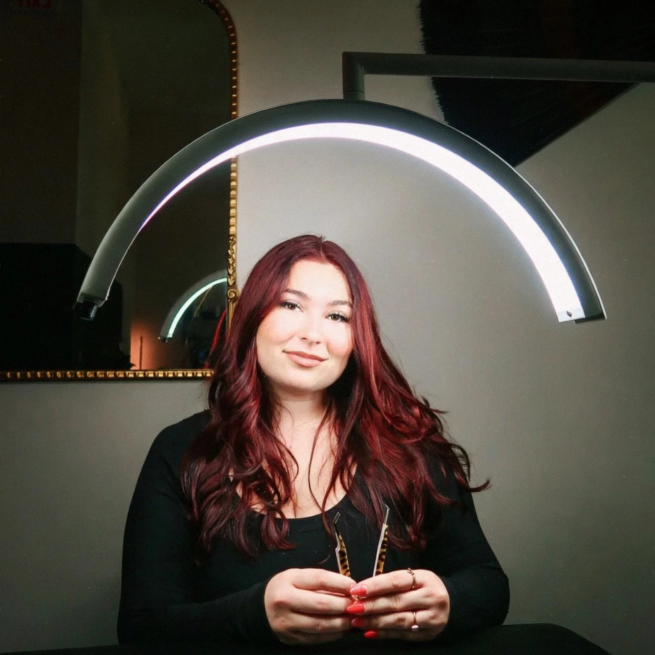 A woman with long, wavy red hair sitting in front of a mirror with a circular light frame. She is smiling and holding her hands together on a black surface.
