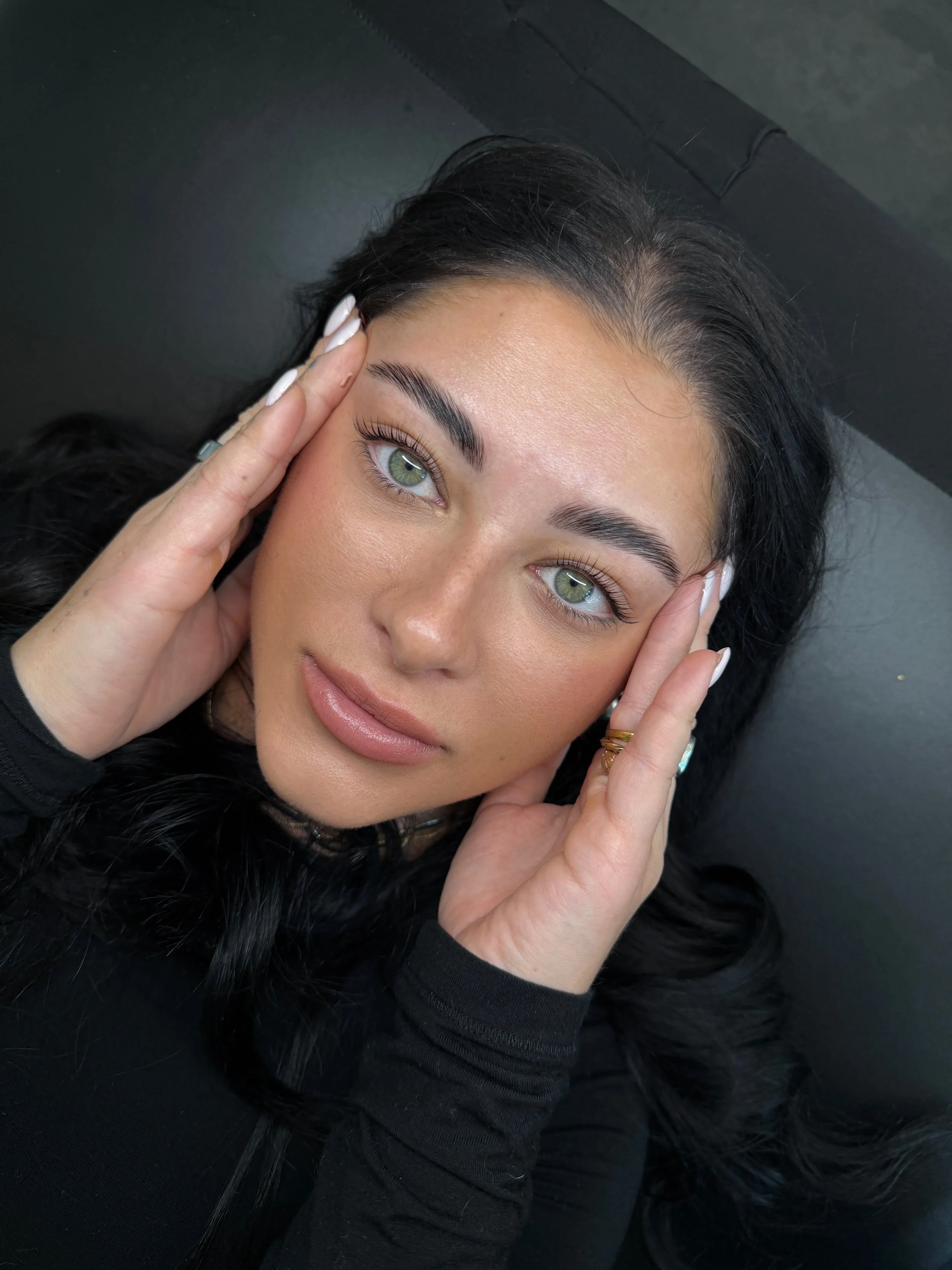 Close-up of a woman with long dark hair and light skin, laying on her back, holding her face with her hands, in front of a dark background, looking directly at the camera.