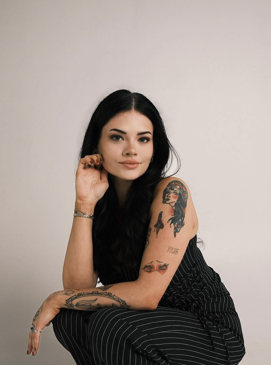 A young woman with black hair, tattoos on her arm, and wearing a black pinstripe dress, sits against a plain light-colored background. She rests her chin on her hand and looks into the camera.