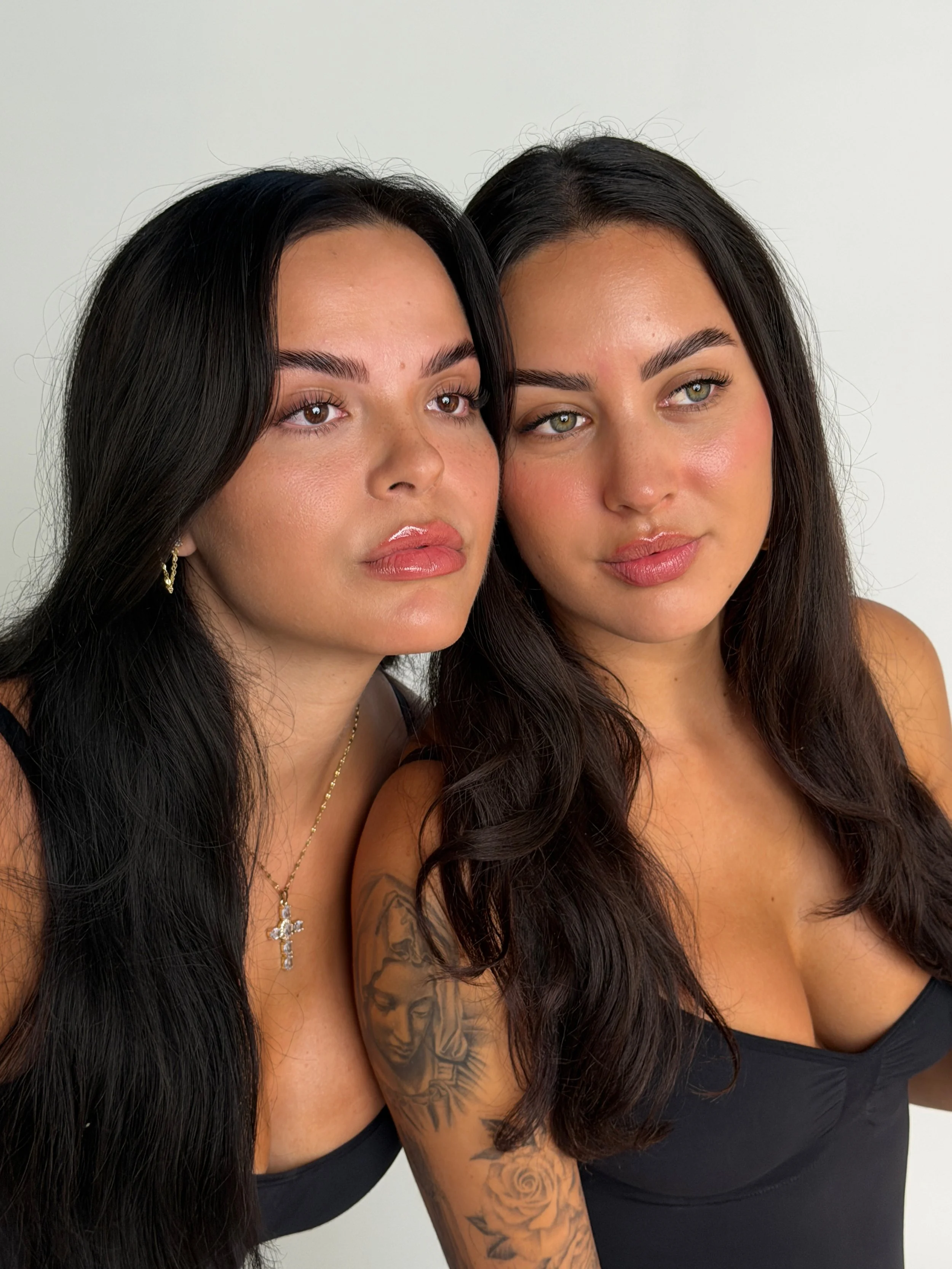 Two women with long dark hair, wearing black tops, standing close together and looking slightly upward. One woman has tattoos on her arm and is wearing jewelry including earrings and a necklace with a cross pendant. The background is plain white.