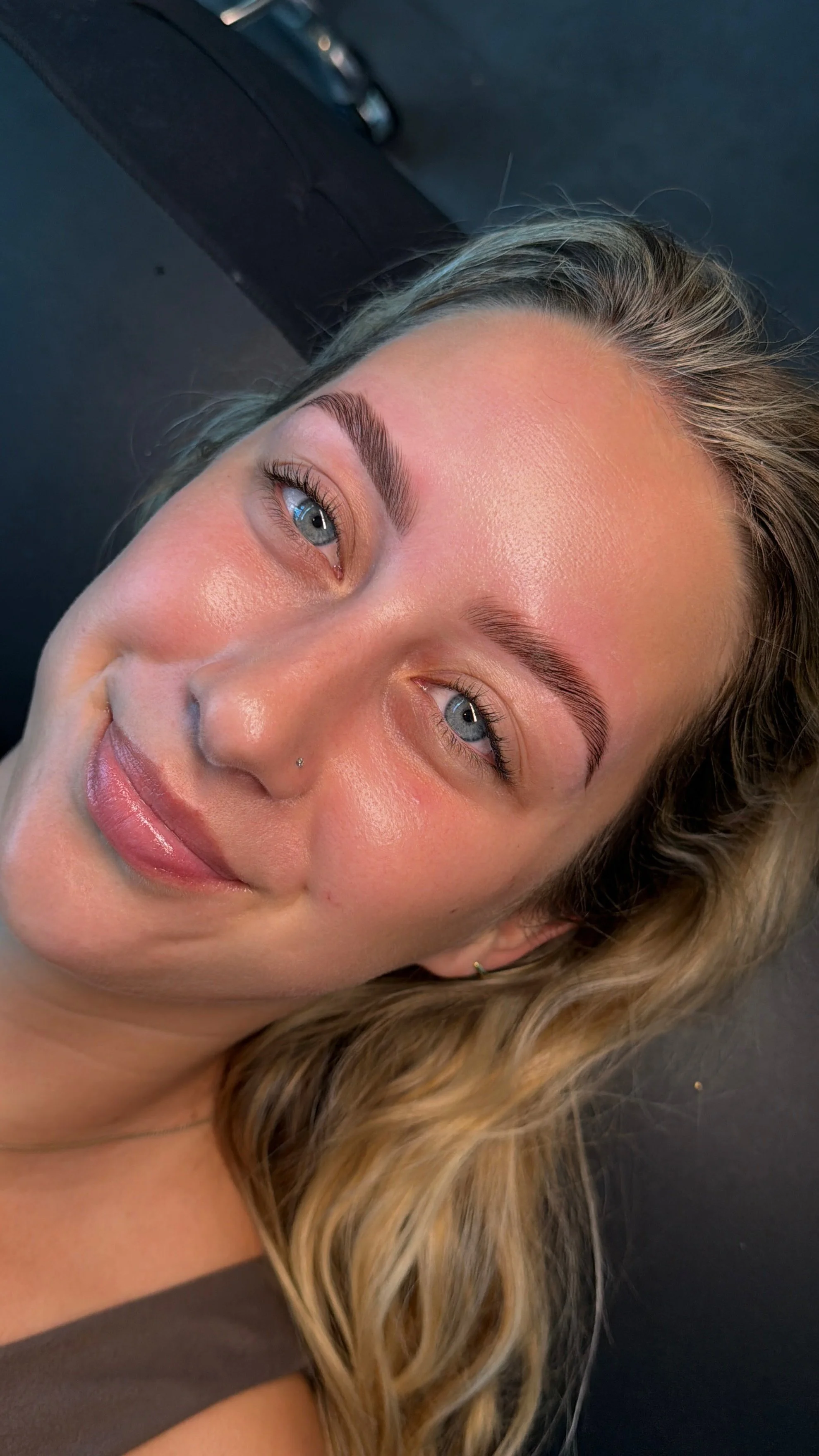 Close-up of a woman with blonde hair, blue eyes, and well-groomed eyebrows, smiling softly, with natural makeup and a small nose piercing.