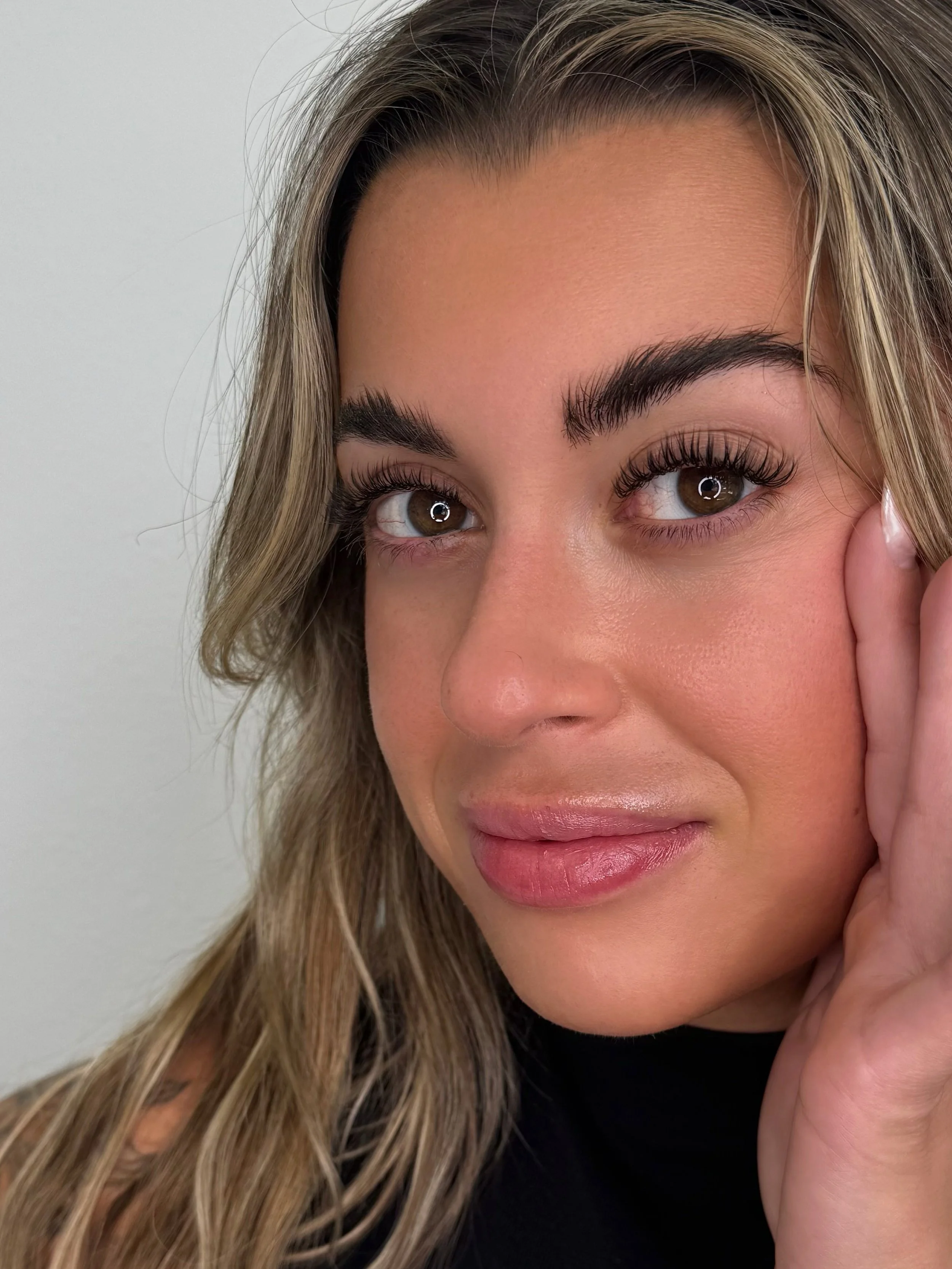 Close-up of a woman's face, showing her left eye, cheek, and part of her lips. She has long, wavy blonde hair, well-groomed eyebrows, and is touching her face gently with her hand.