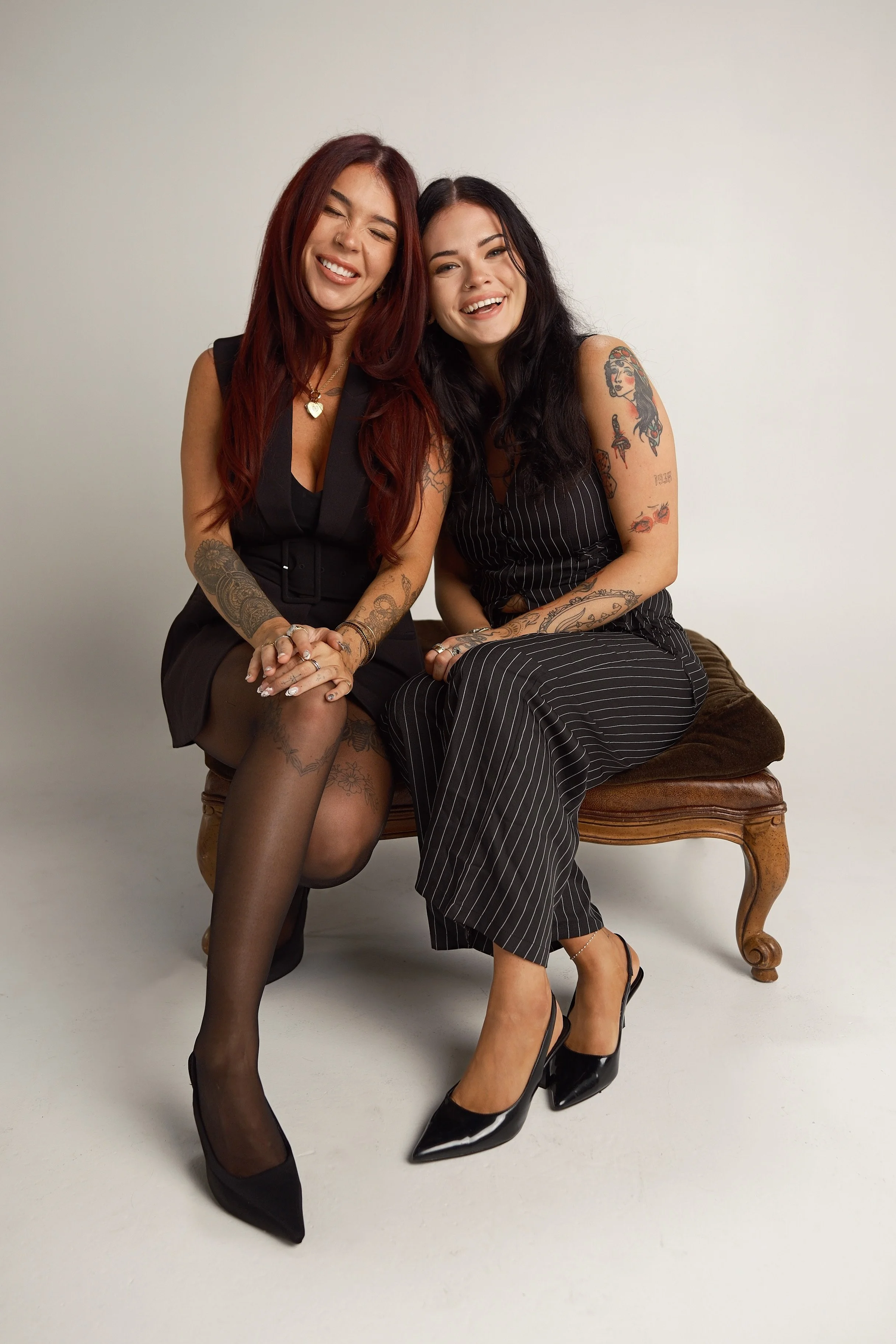 Two women sitting on a vintage wooden bench against a plain white wall, smiling and laughing. Both are dressed in black outfits with tattoos visible on their arms, and wearing black high heels. One woman has long red hair, the other has long black hair.