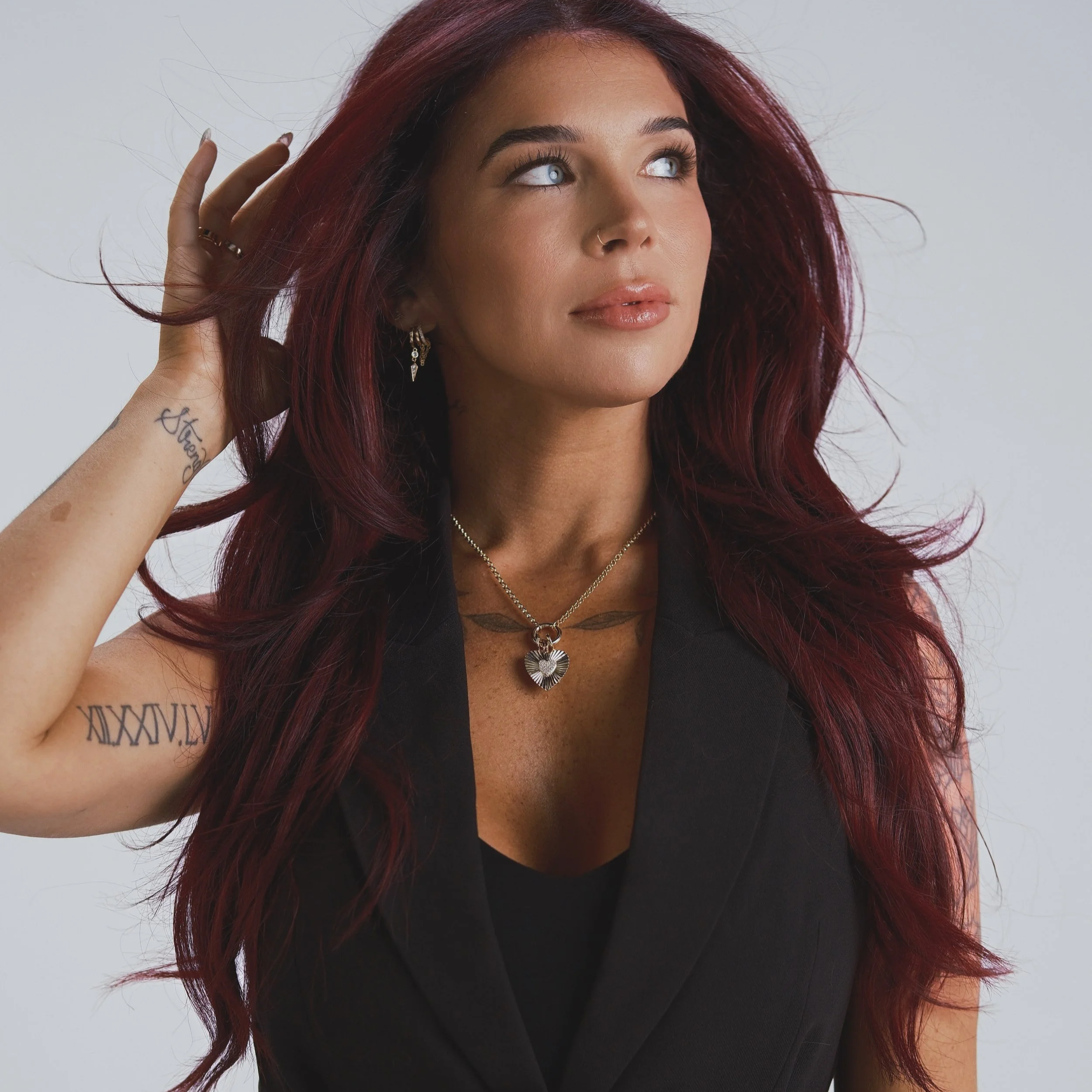 A woman with long, wavy red hair looking upward and to the right, touching her hair with her left hand, wearing jewelry including earrings and a necklace, with tattoos on her left arm, against a plain, light background.