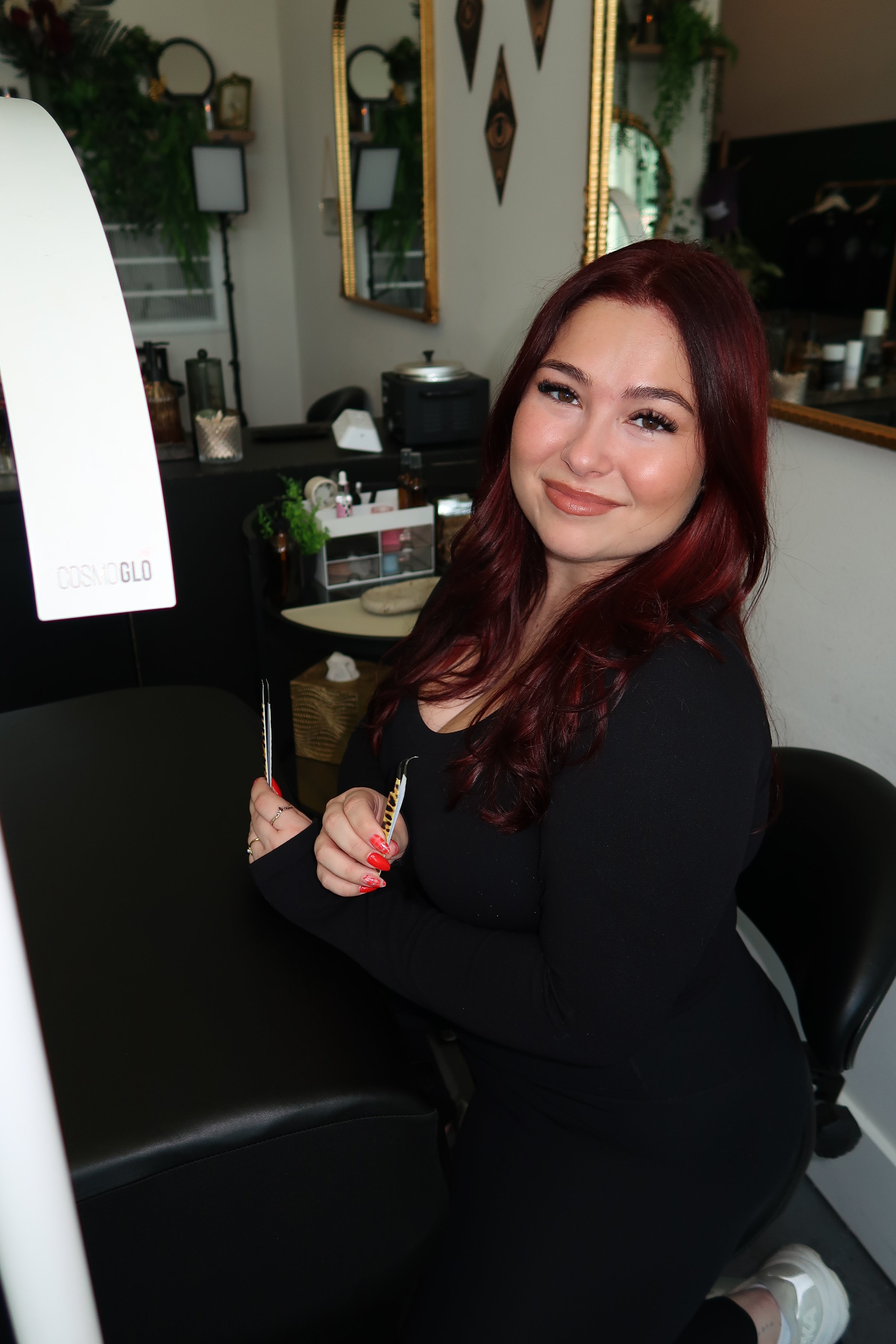 A young woman with long, wavy red hair and light makeup sitting in a salon or beauty studio, smiling at the camera and holding a small makeup brush.