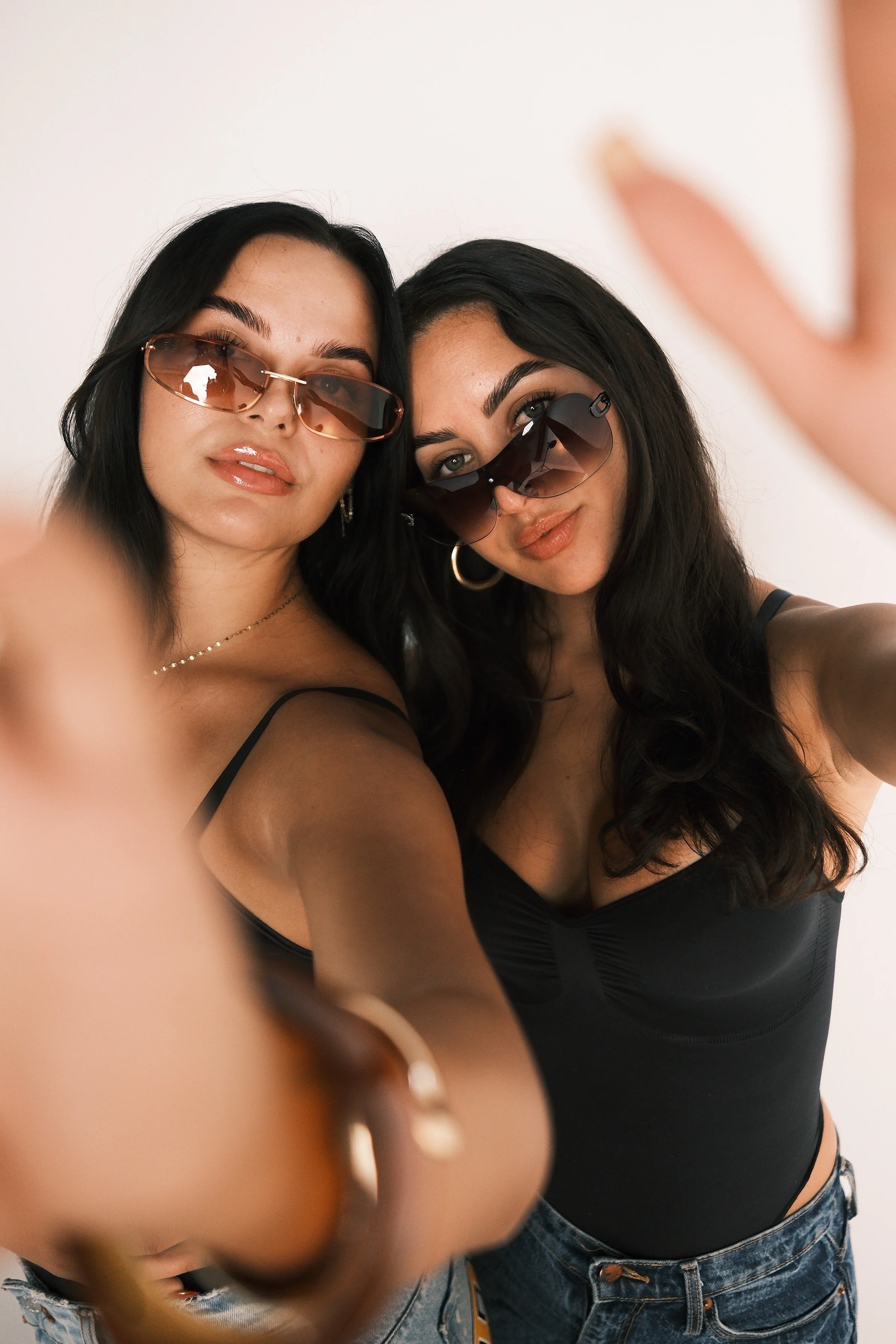 Two women taking a selfie, both wearing sunglasses and black tops, with one woman with dark hair and the other with black hair, against a plain background.