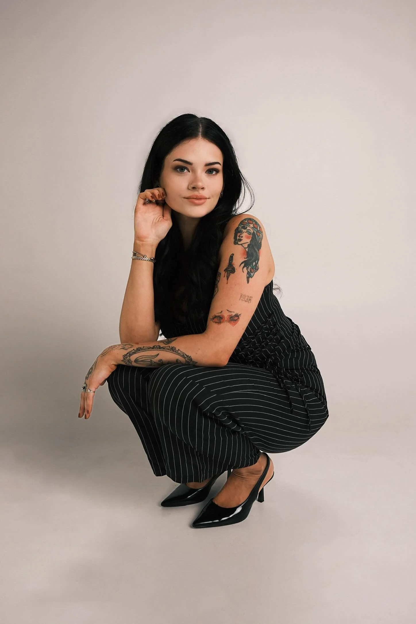 A woman with black hair and tattoos, dressed in a black and white striped outfit, poses against a plain light-colored background, wearing black high heels.