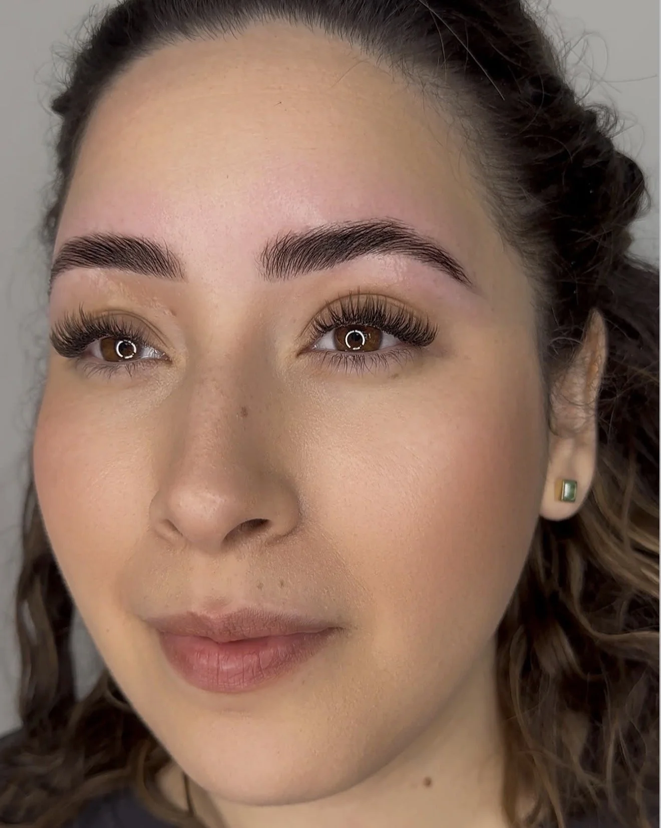Close-up of a woman's face showing her brown eyes, well-groomed eyebrows, and makeup. She has curly brown hair and is wearing a small earring with a green stone.