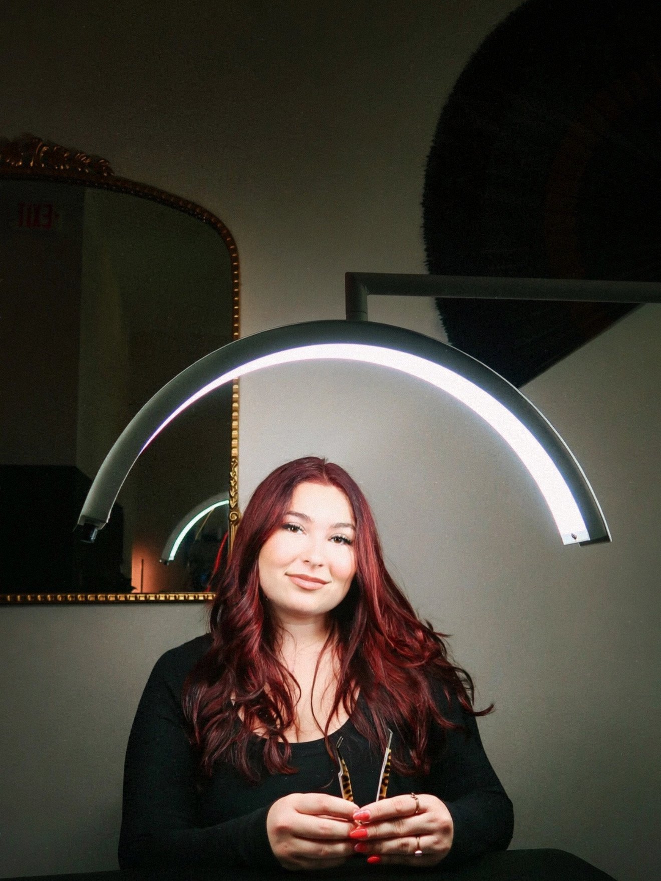 A woman with long red hair and a black top sitting in front of a mirror with a modern, curved, illuminated ring light above her head, in a room with gray walls and a black spherical pendant light in the background.