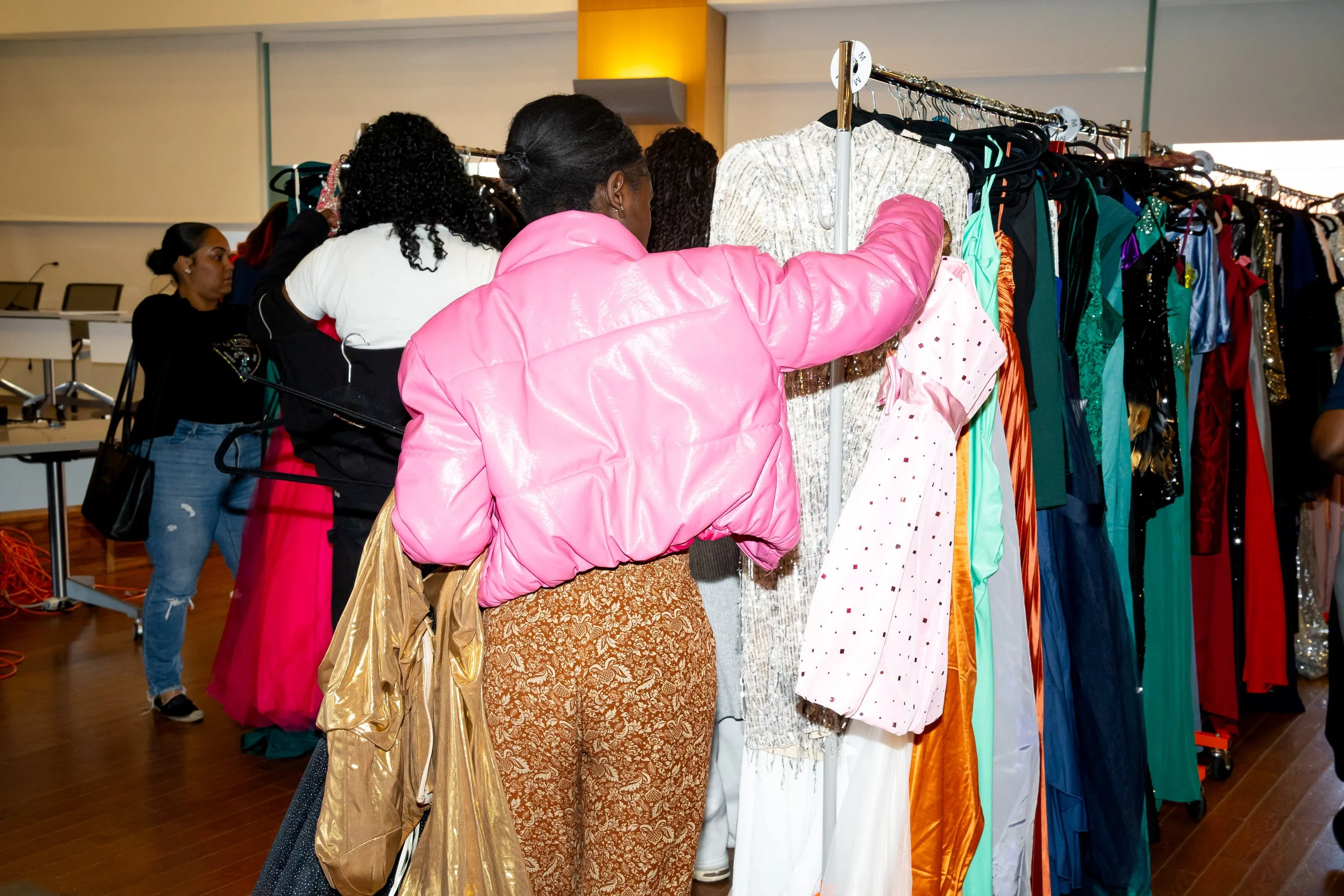 Boston Public Schools student in bright pink jacket and patterned pants looks through a rack of formal dresses.