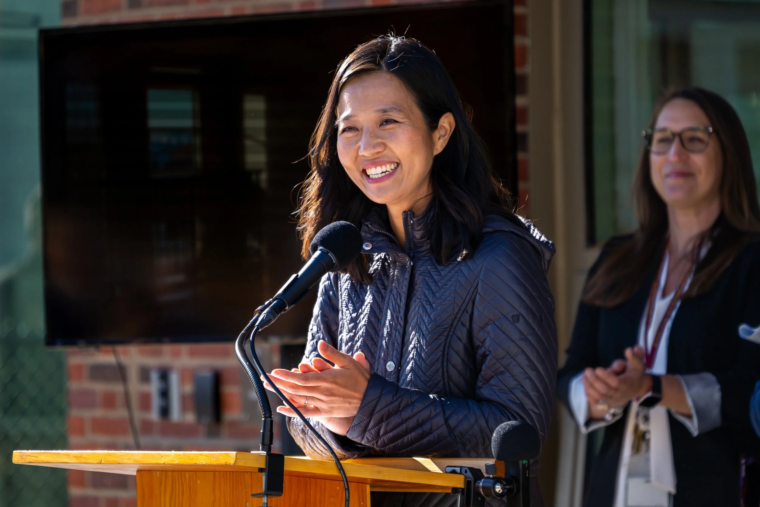 City of Boston Mayor Michelle Wu smiles and claps her hands while standing at a podium.