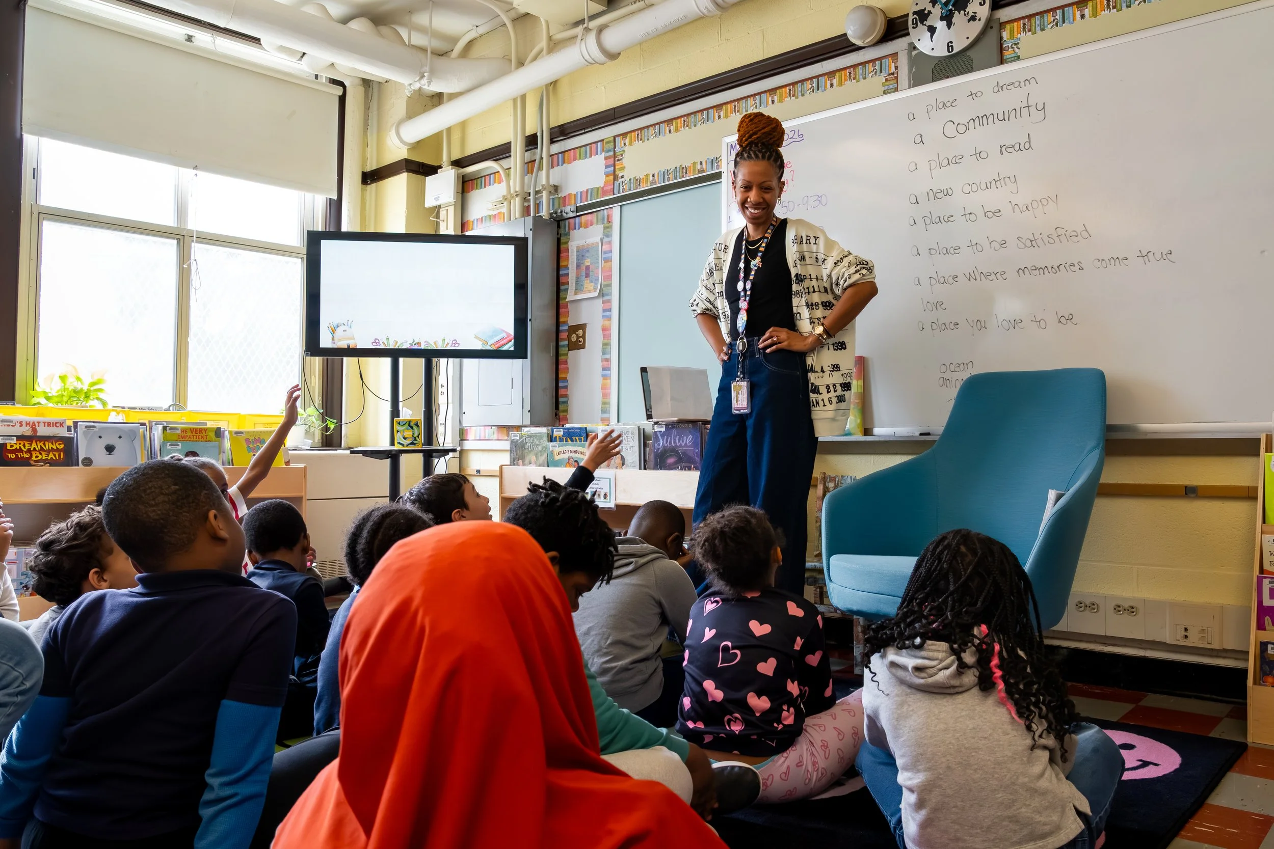 Boston Public Schools librarian Kadian Simmonds smiles and stands in front of a group of students seated on a rug.