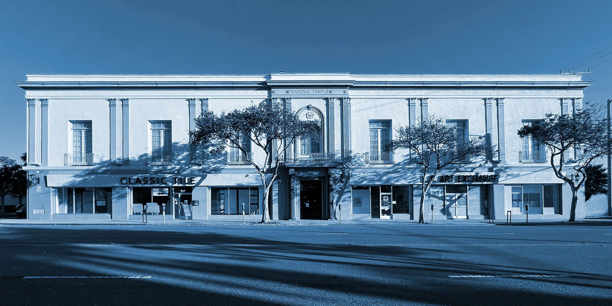 A classic two-story white building with architectural columns, large windows, and a balcony above the entrance, with store signs reading "Classic Tile" and "Art Exchange" on the ground floor and trees in front.