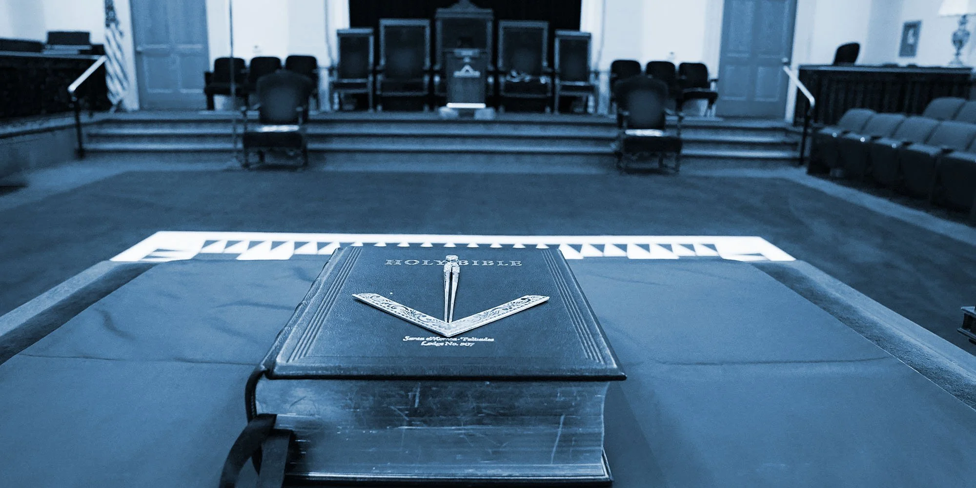 A Bible resting on a table in a church with a sign on the floor that says 'Fellowship'. The church has a stage with chairs and a podium in the background.
