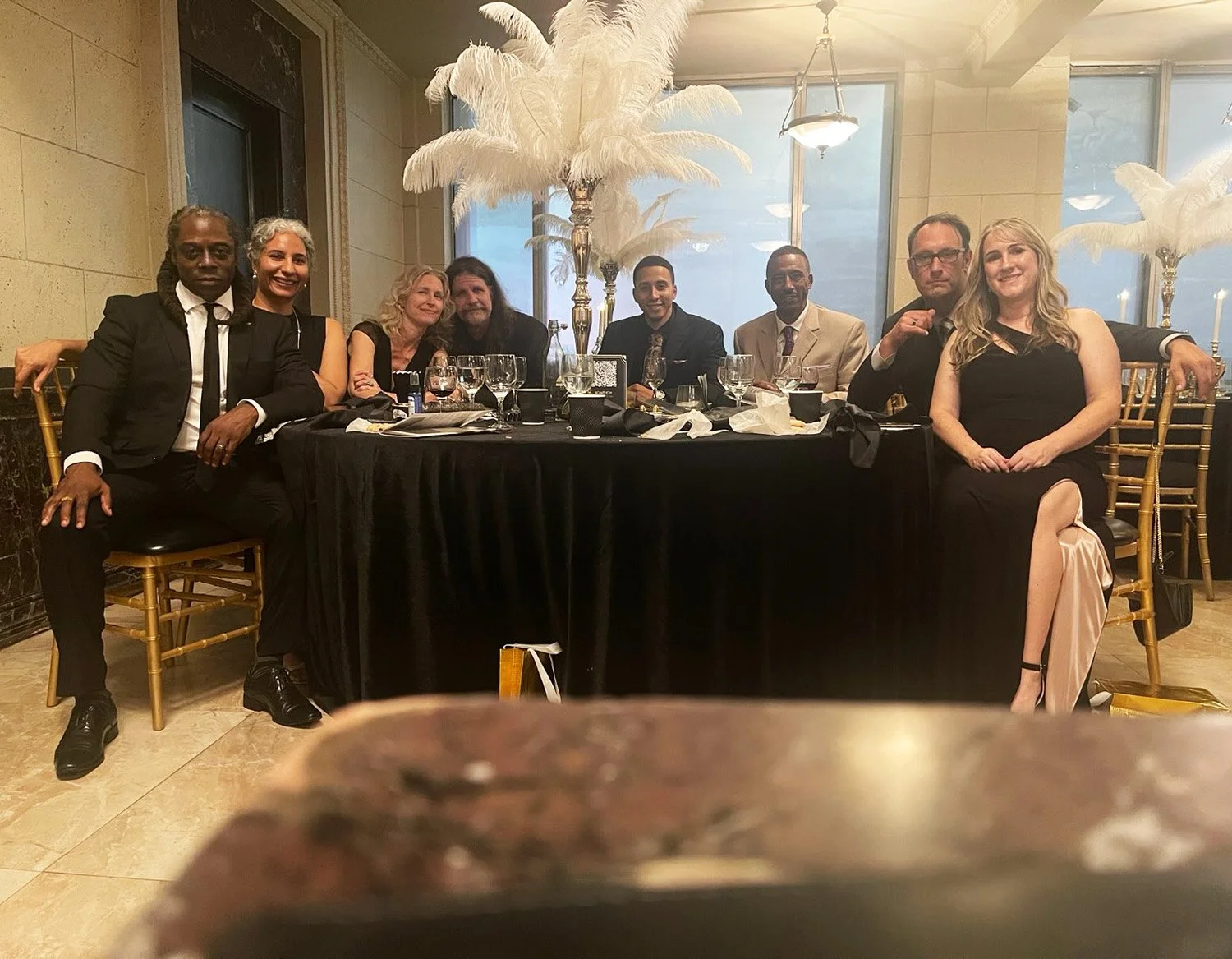 Freemasons and their companions around a dining table at a formal event. The table is set with glasses and plates, and decorated with large white feather centerpieces. The background shows a large window and elegant decor.