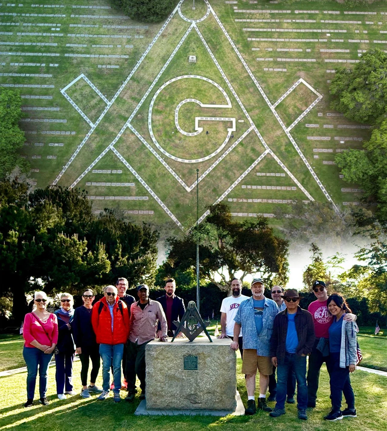 A group of eleven people standing outdoors in a park, smiling at the camera. Behind them is a monument with a large square and a compass and square symbol, and above them, an aerial view of a large memorial area with a design of a triangle and a letter G in the center, made of white lines on the grass, surrounded by trees.