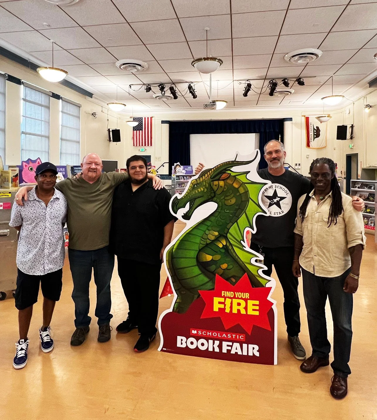 Group of five people standing together at a book fair, posing behind a large green dragon cutout with the text 'Find Your Fire' and 'Scholastic Book Fair'.