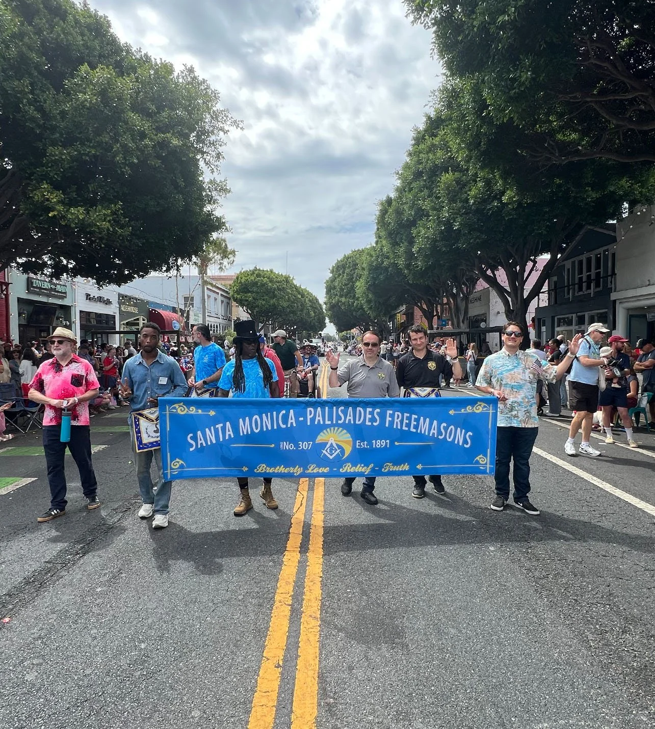 A parade on a city street featuring a group of men holding a blue banner for Santa Monica-Palisades Freemasons, with people watching on the sides. The street is lined with trees and storefronts, with a cloudy sky overhead.