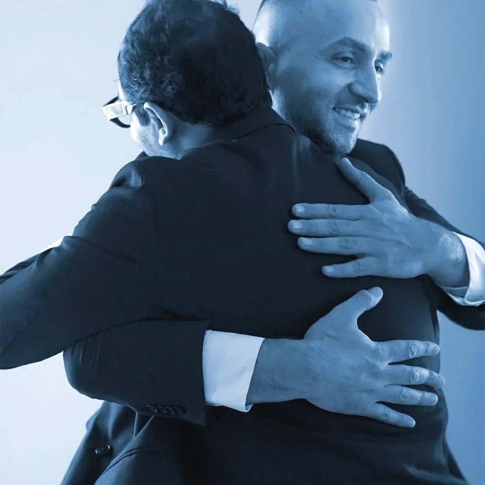 Two men in formal suits are embracing each other warmly, smiling, in a friendly hug.
