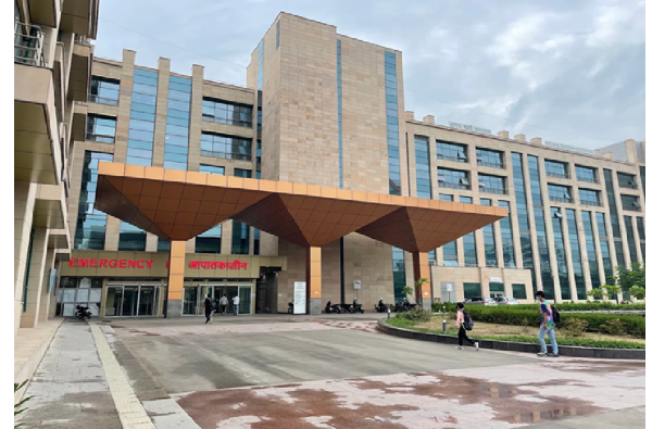 Modern hospital building with a large, brown, canopy entrance. The building has multiple floors with glass windows and an emergency entrance marked in red. People are walking in and out of the hospital on a cloudy day.