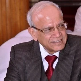 An elderly man with white hair and glasses wearing a dark suit, white shirt, and red tie, smiling in a formal setting.