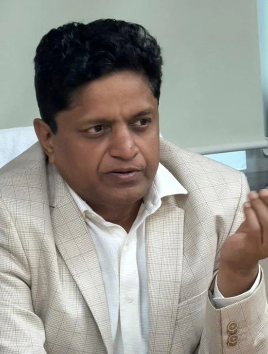 A man with dark hair in a beige plaid blazer and white shirt, seated at a desk, gesturing with his right hand, and speaking or explaining something.