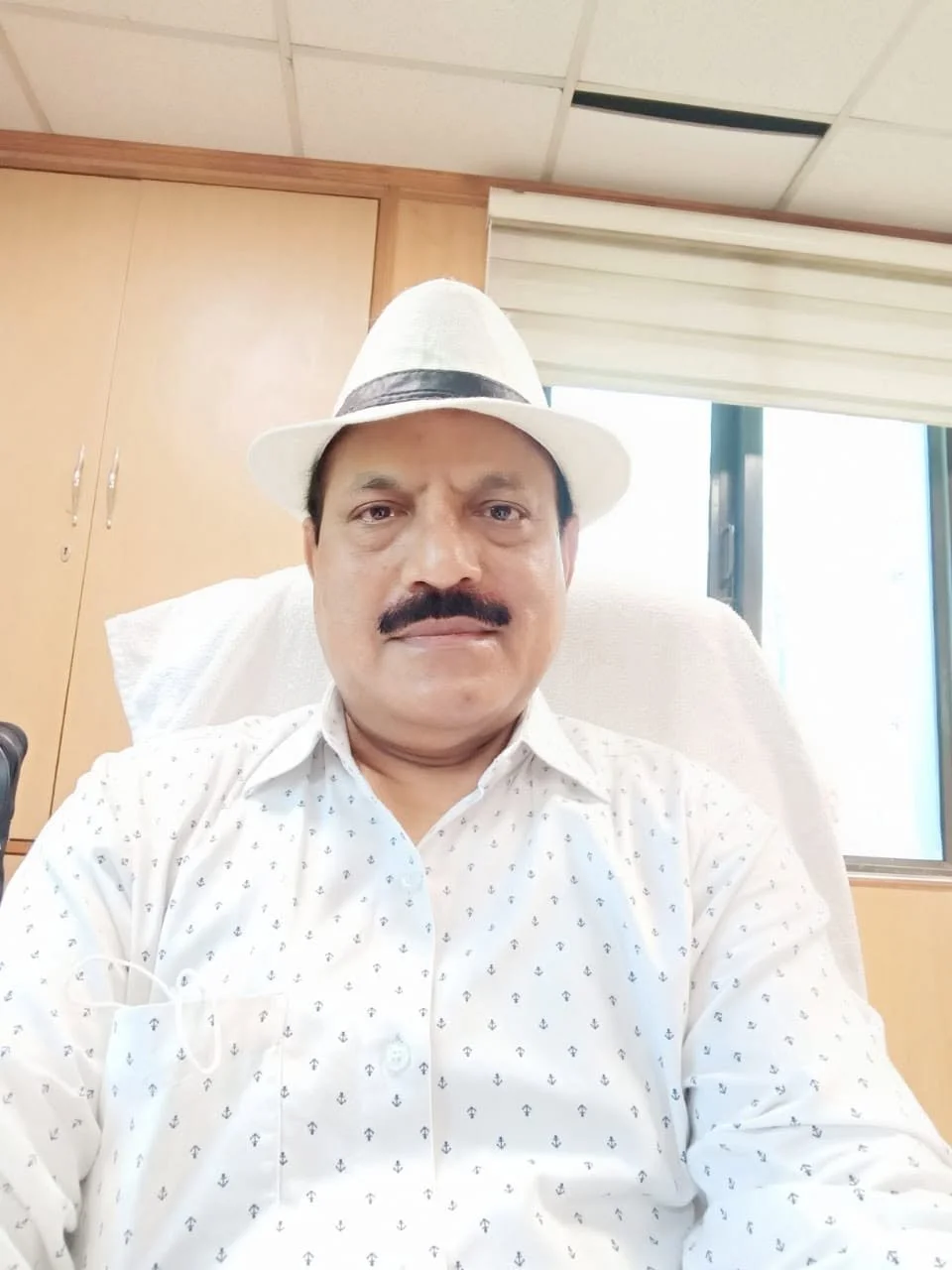 A man with a mustache wearing a white hat and a white shirt with small dark patterns, sitting in an office, with a window and wooden cabinets in the background.