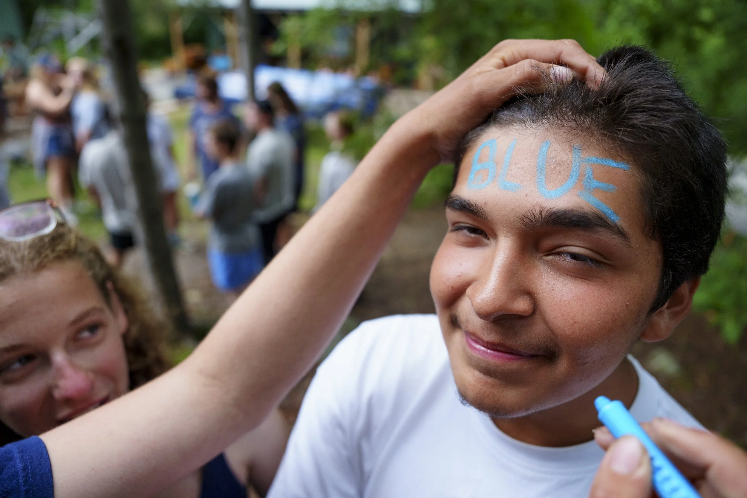 Linden Wylie writes the text Blue on Mario Lopez' face in blue face crayon to identify what team he plays for in the Camp Olympic Games hosted by Camp Temagami at the conclusion of the summer of wilderness canoe trips, August 8, 2026