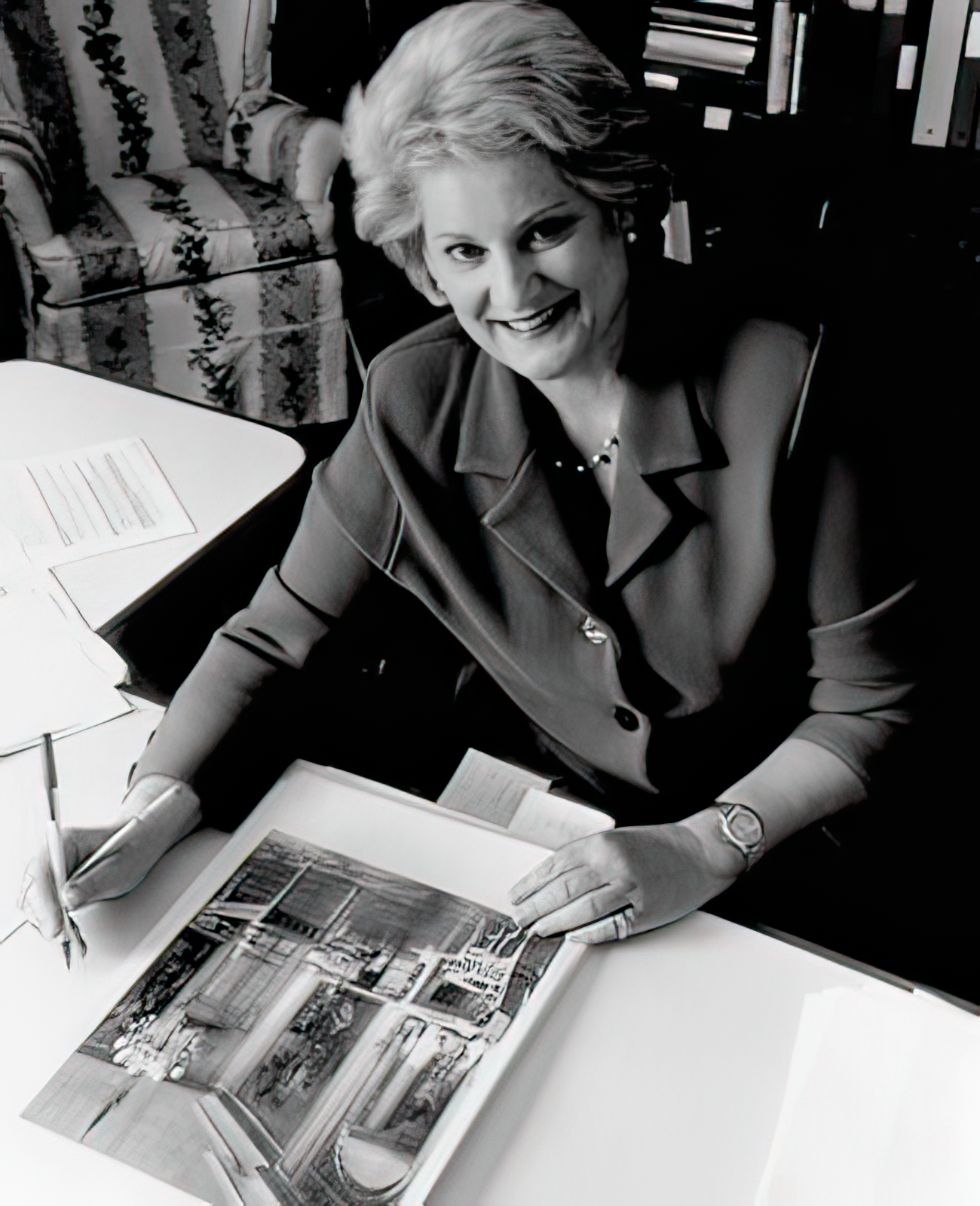 Mary Frick Symington Sanger is sitting at a table with a large open photo album, looking up at the camera and smiling. She appears to be reviewing photographs or artwork, with various papers and a small umbrella on the table.
