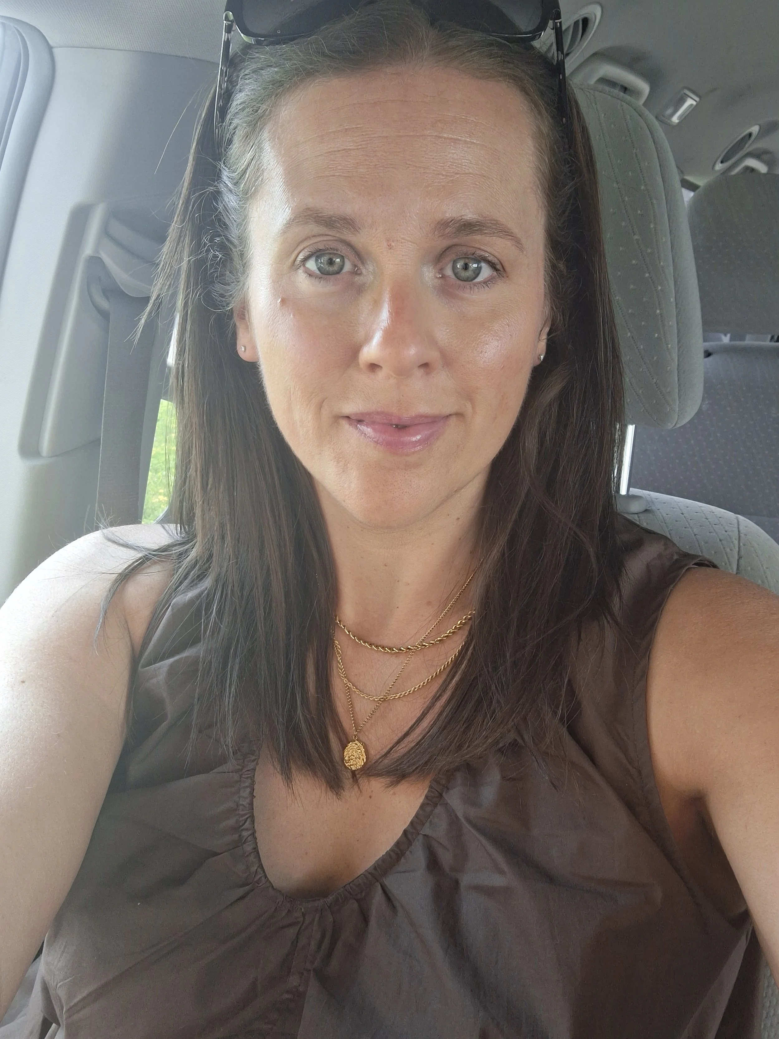 A woman with blue eyes, long dark hair, wearing sunglasses, gold jewelry, and a dark brown sleeveless top, sitting inside a vehicle.