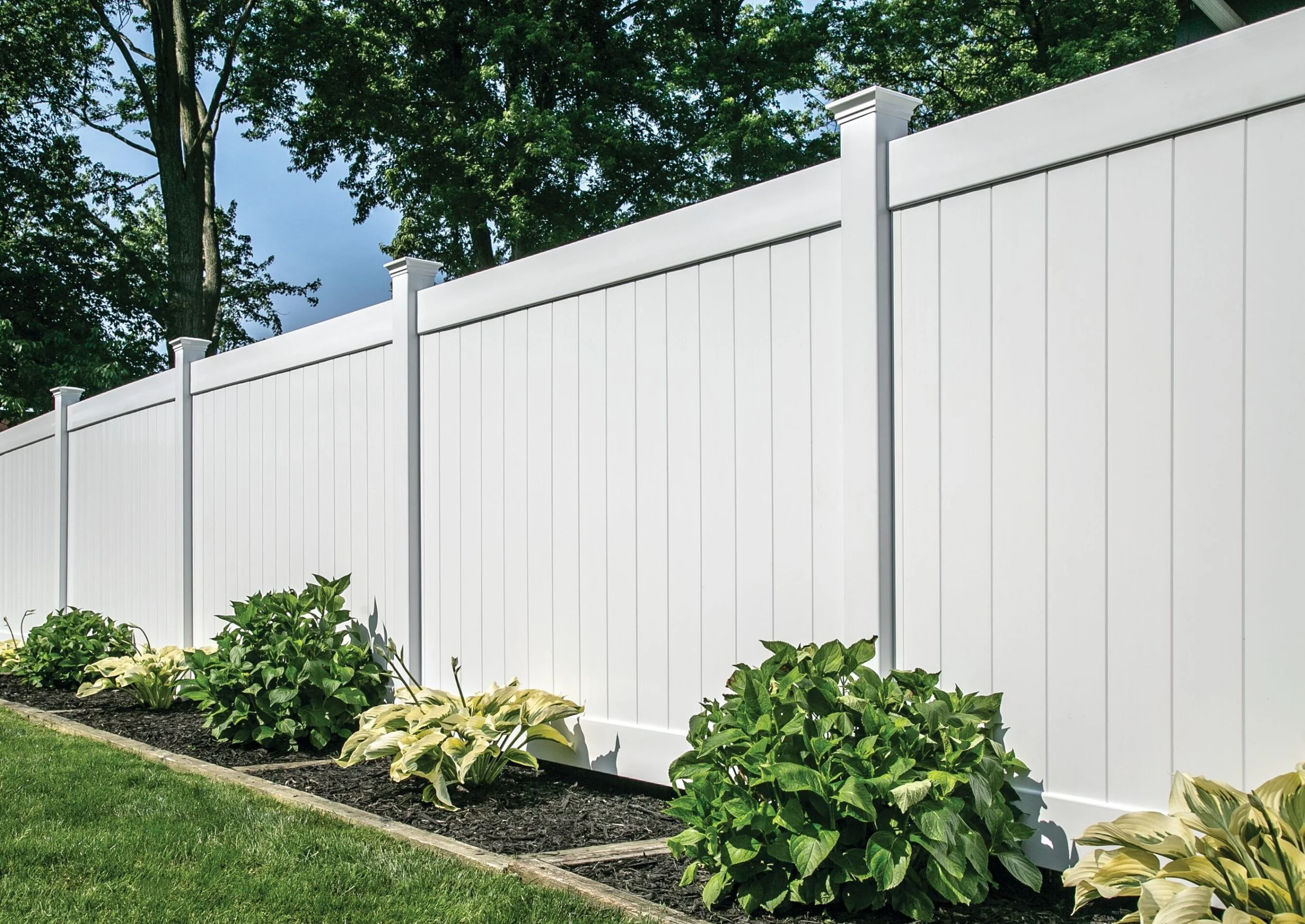 A white vinyl privacy fence with decorative post caps in a backyard garden with green bushes and a lawn.