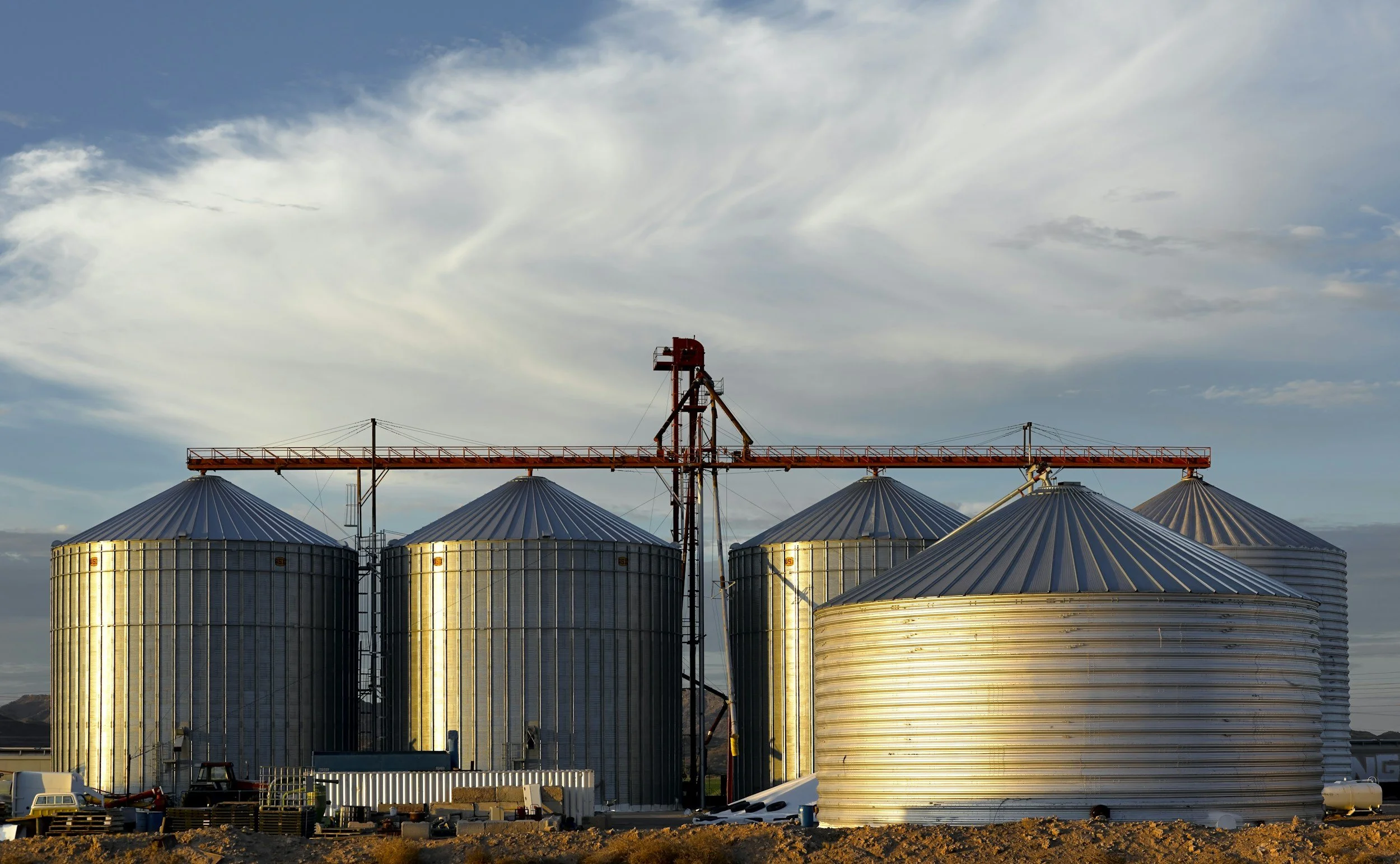 Large metallic grain silos with a red crane structure above them, set against a cloudy sky.