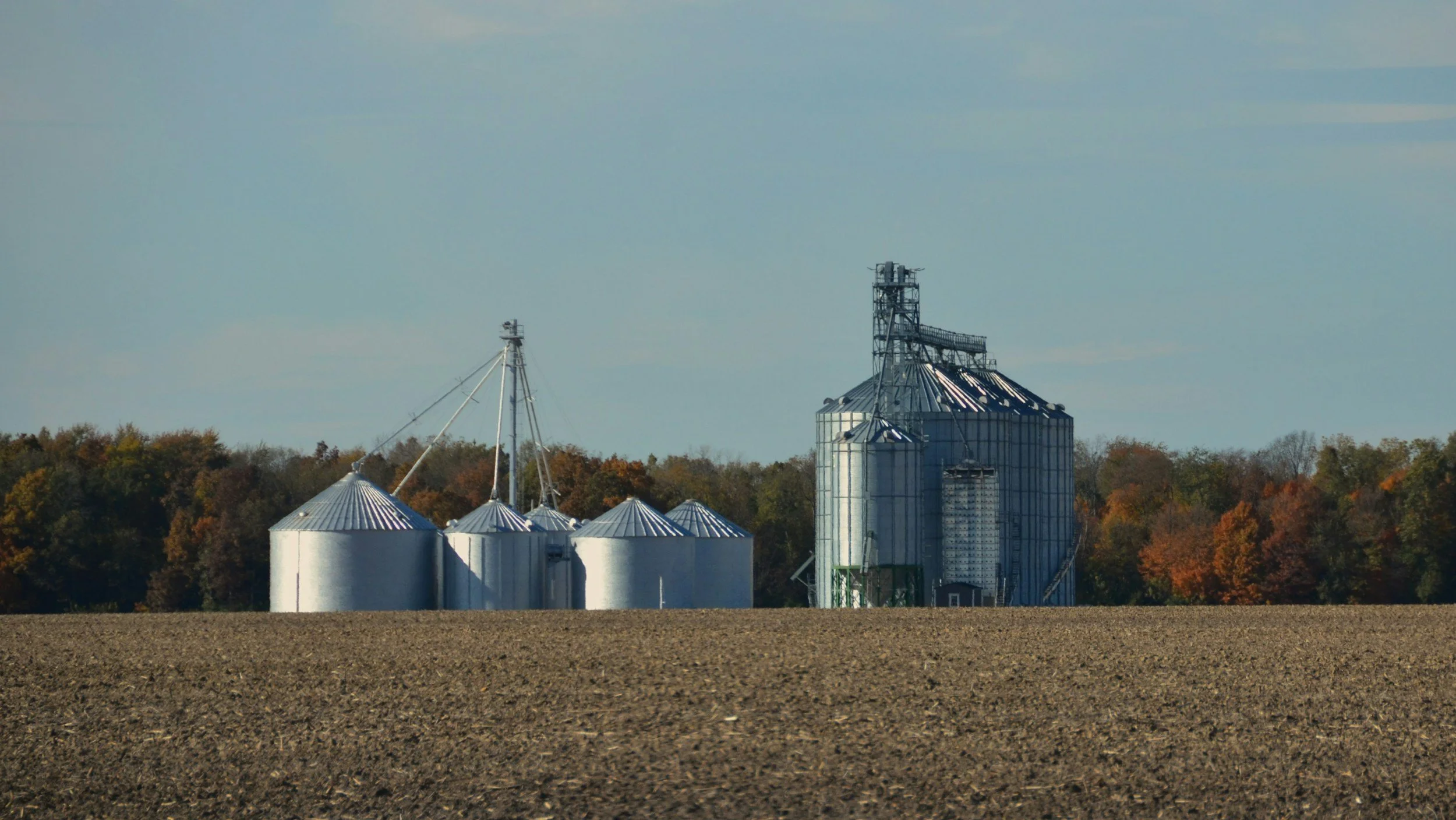 Agricultural farm with metal silos and a grain elevator against a background of trees with autumn foliage and a partly cloudy sky.