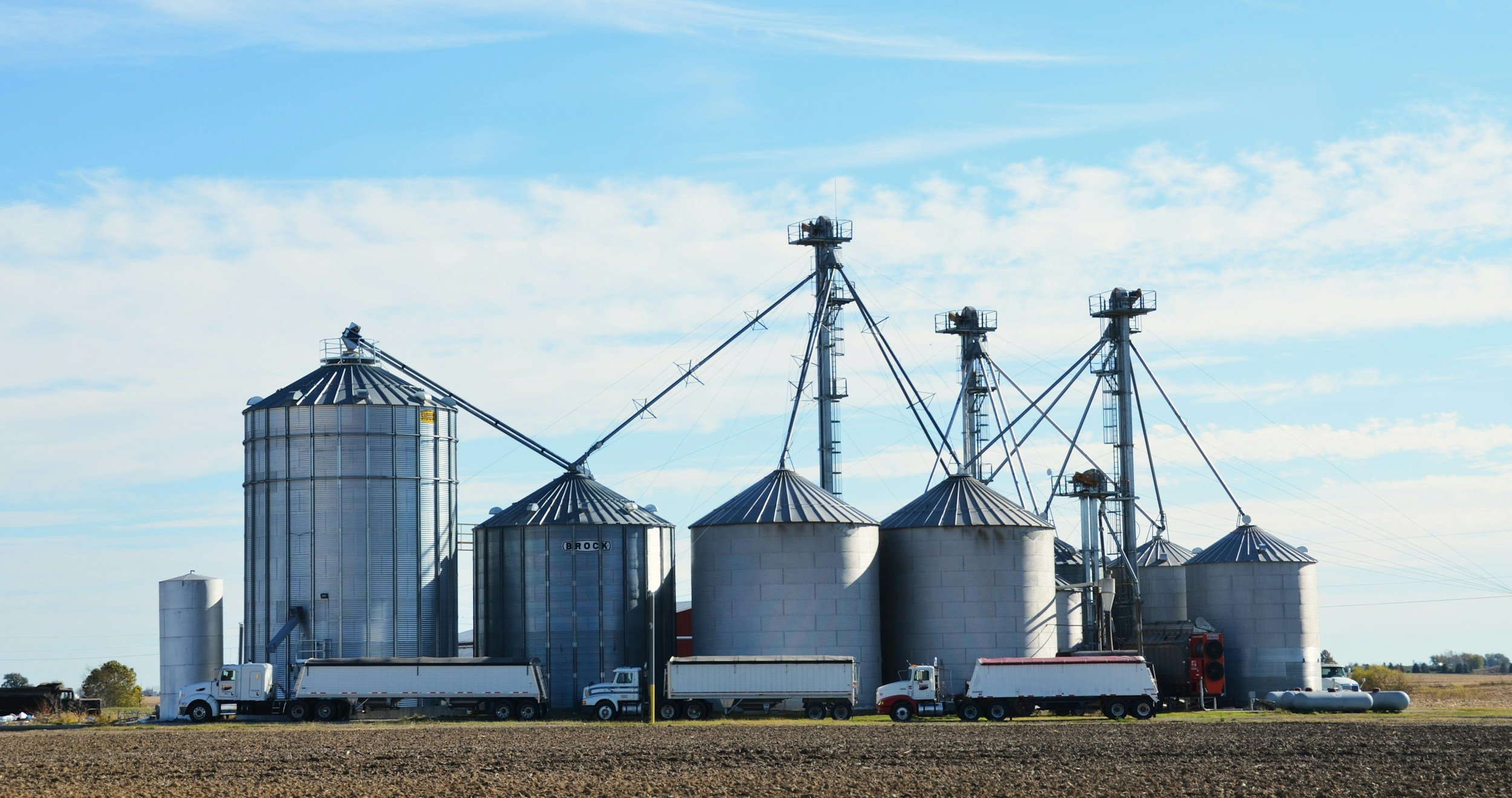 Silage storage silos and grain bins in a rural farm area with trucks parked in front, under a partly cloudy sky.