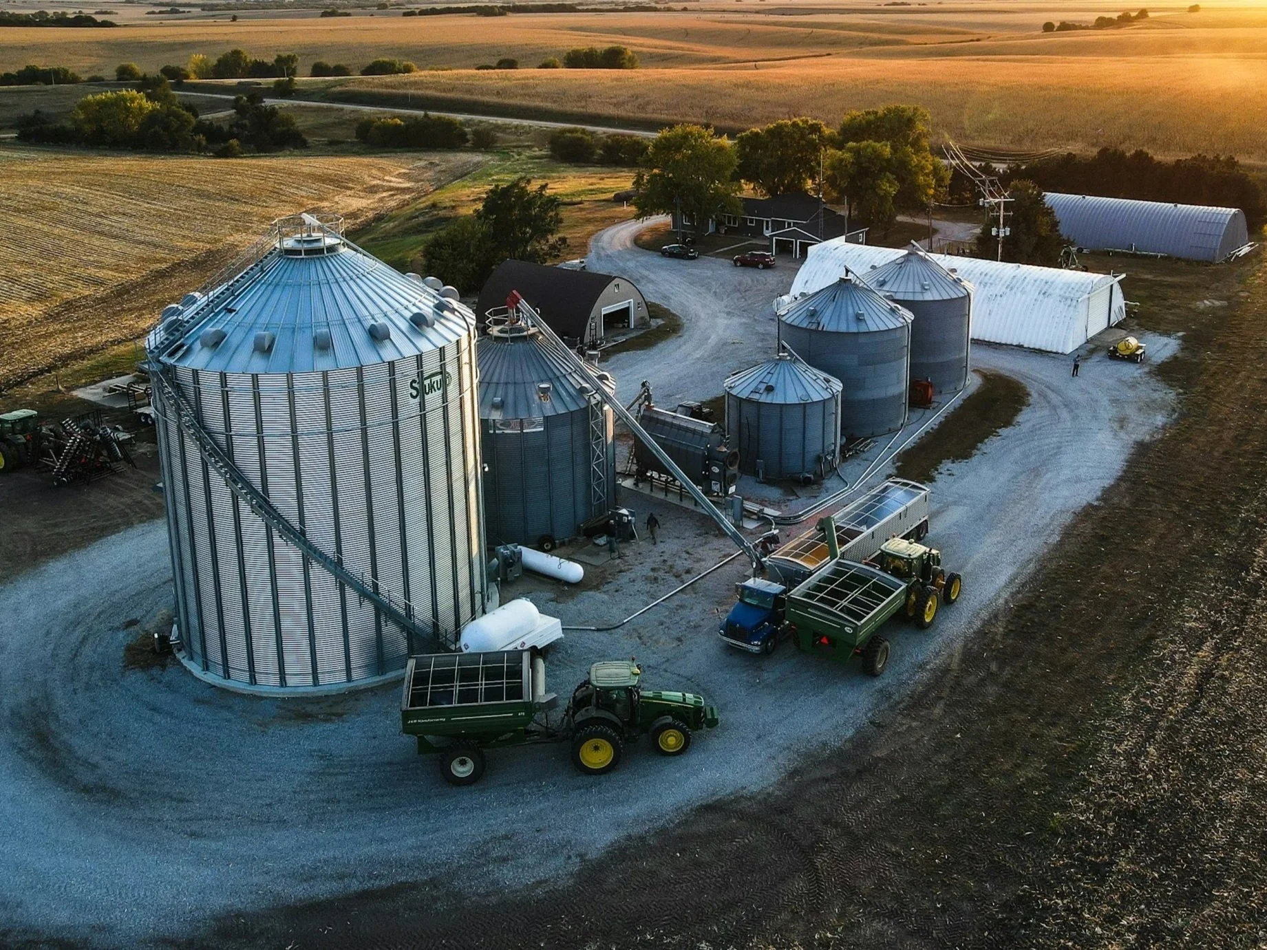 Aerial view of a farm with multiple large metal grain silos, storage buildings, farm equipment, and vehicles, surrounded by fields and trees at sunset.