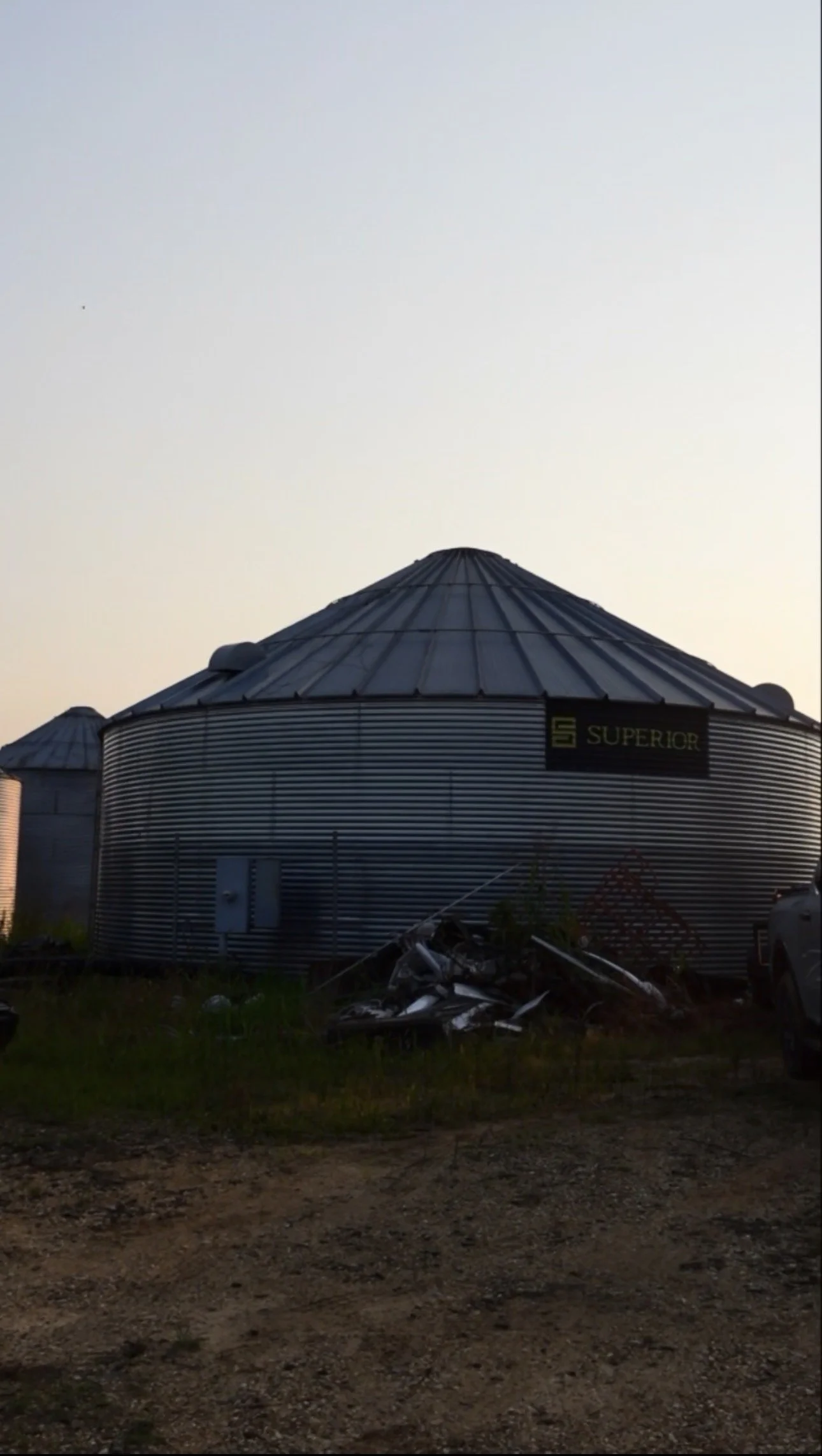 A large metal grain silo with the word 'SUPERIOR' on it, situated outdoors at dusk, with some debris and grass in the foreground.