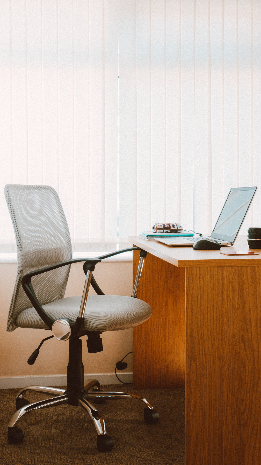An empty office desk with a laptop, mouse, notebooks, a lunchbox, a camera, and a coffee mug, next to an office chair, in front of a window with closed vertical blinds.