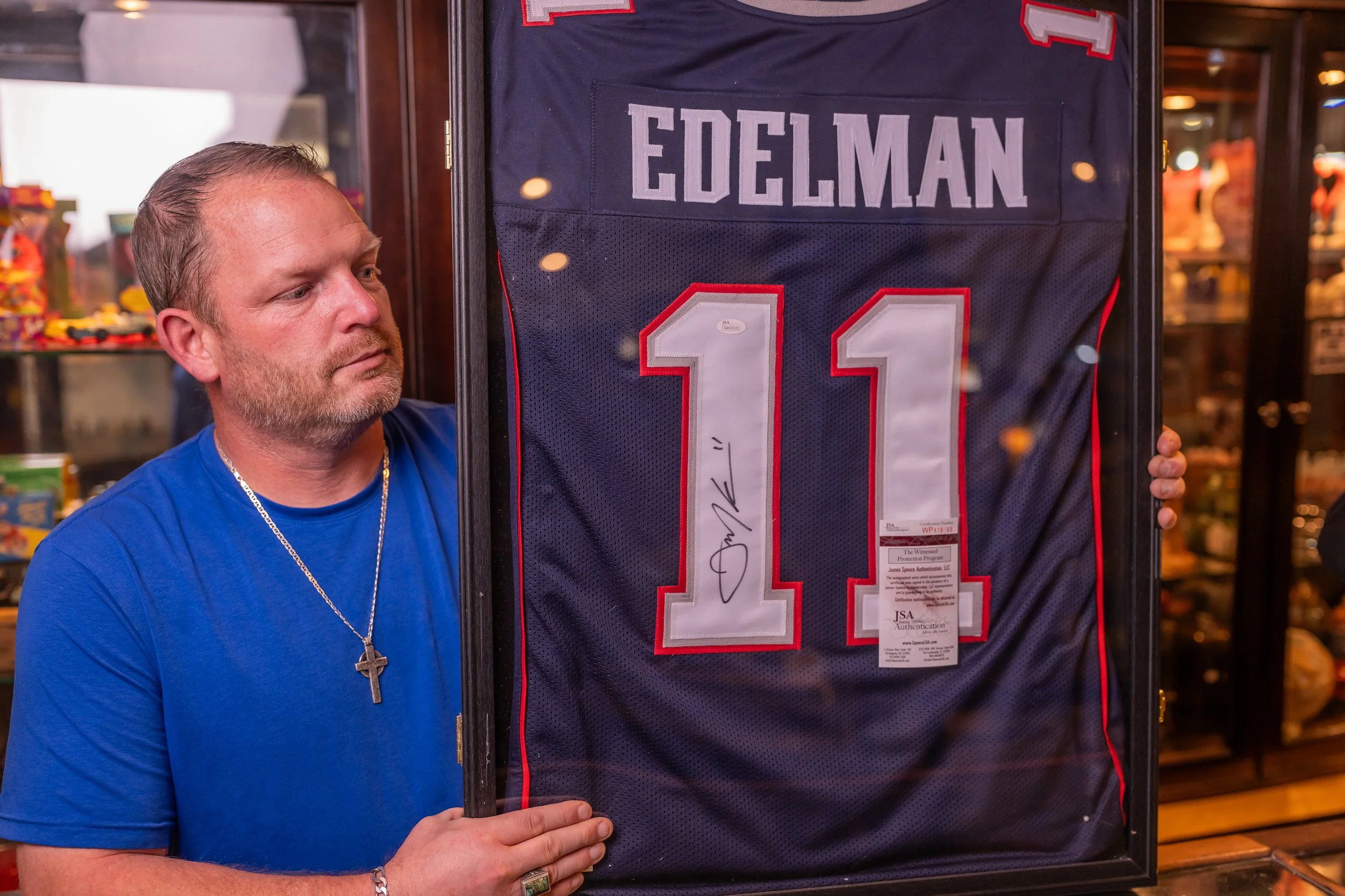 A man with short blonde hair, wearing a blue t-shirt, holds a framed sports jersey with the name "Edelman" and the number 11. The jersey is signed and displayed behind glass.