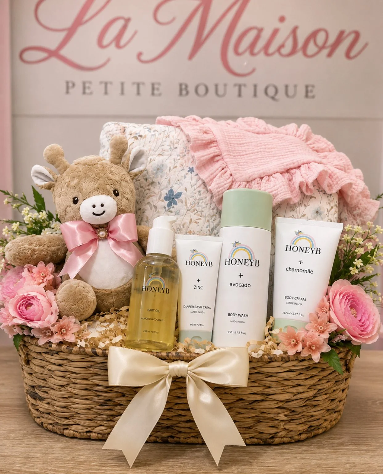 Gift basket with a plush giraffe toy, skincare products, pink and floral fabric, flowers, and a white ribbon, in front of a pink sign that reads 'La Maison Petite Boutique'.