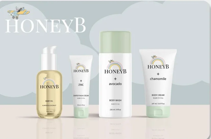 Collection of HoneyB skincare products including baby oil, diaper rash cream with zinc, avocado body wash, and chamomile body cream, arranged on a light background.