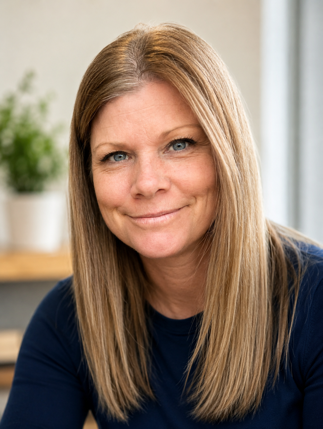 Close-up of a woman with long blonde hair, blue eyes, smiling, wearing a dark blue top, with a blurred plant in the background.