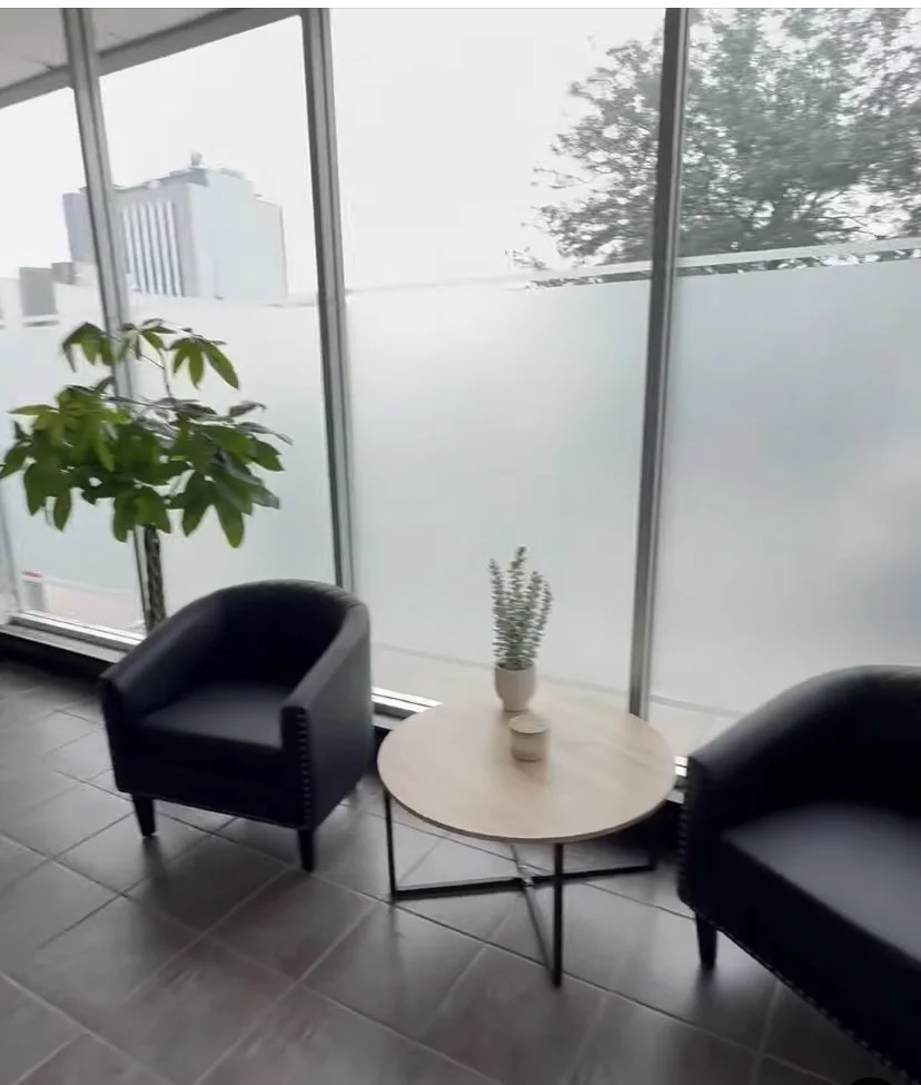 Indoor view of a modern seating area with two black armchairs, a round wooden coffee table with a potted plant, and large frosted glass windows showing an urban cityscape outside.