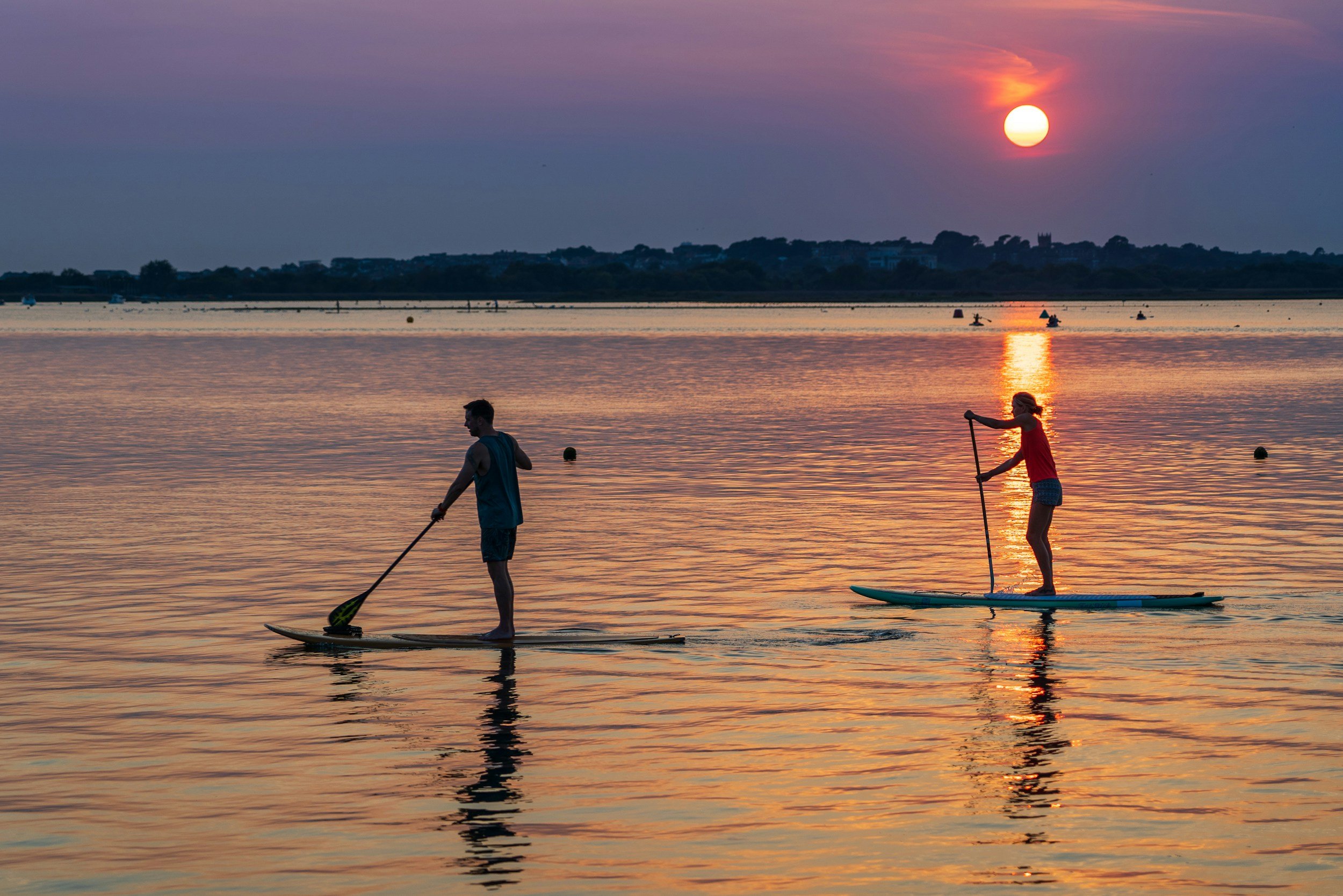 Two people paddleboarding on calm water during sunset, with the sun low on the horizon and pink and purple sky, in a scenic outdoor setting.