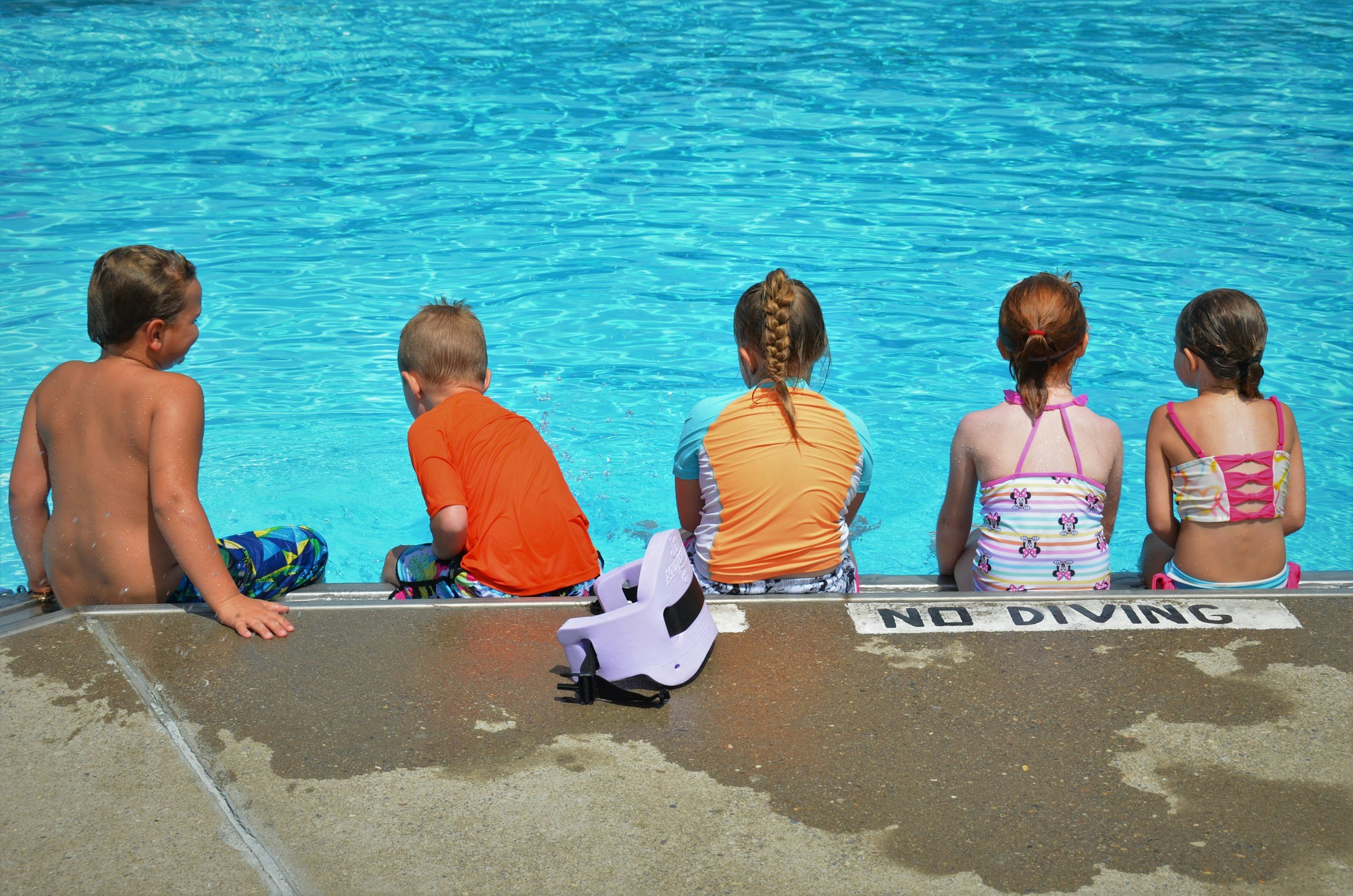 Five children sitting at the edge of a swimming pool, viewed from behind, with water in the background. The children are wearing swimsuits and summer clothes, with a white sign on the poolside reading 'NO DIVING.'